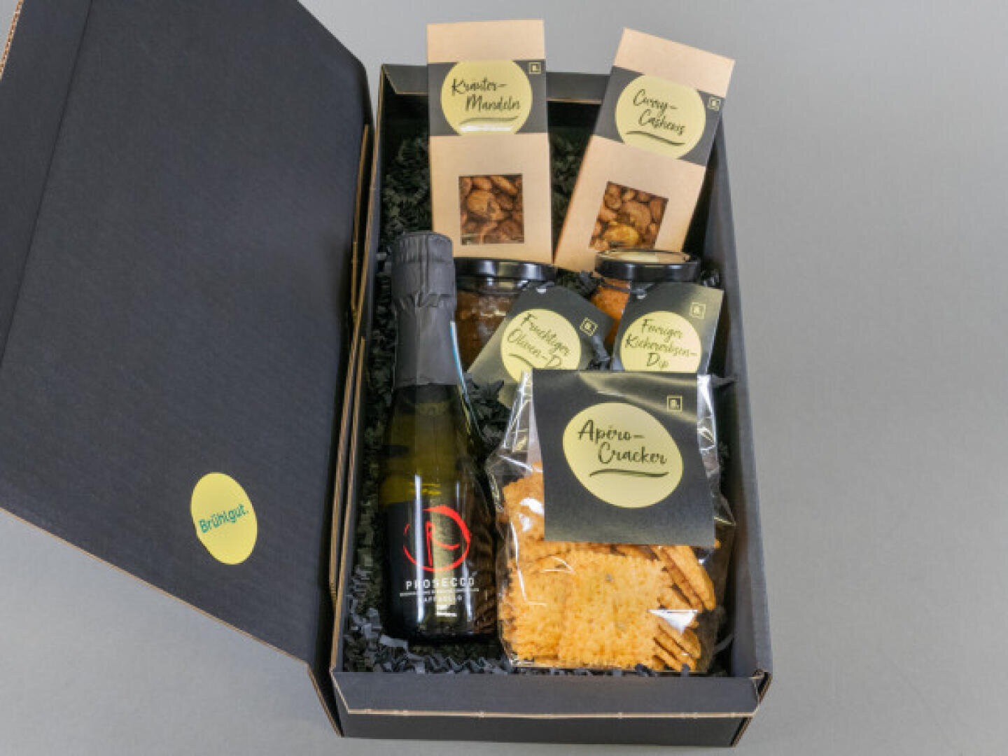 A gift box with a small bottle of Prosecco, crackers, two jars of dip and three packets of snacks, all neatly arranged on black crinkle paper.