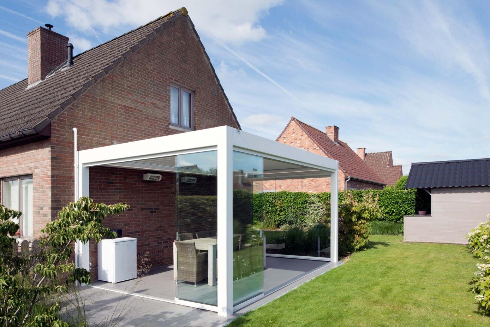 Eine moderne gläserne Terrassenüberdachung mit weißem Rahmen ist an ein Backsteinhaus angebaut, in dem ein Tisch und Stühle stehen. Die Überdachung ist von grünem Rasen und einem Garten umgeben, über dem sich ein strahlend blauer Himmel spannt.