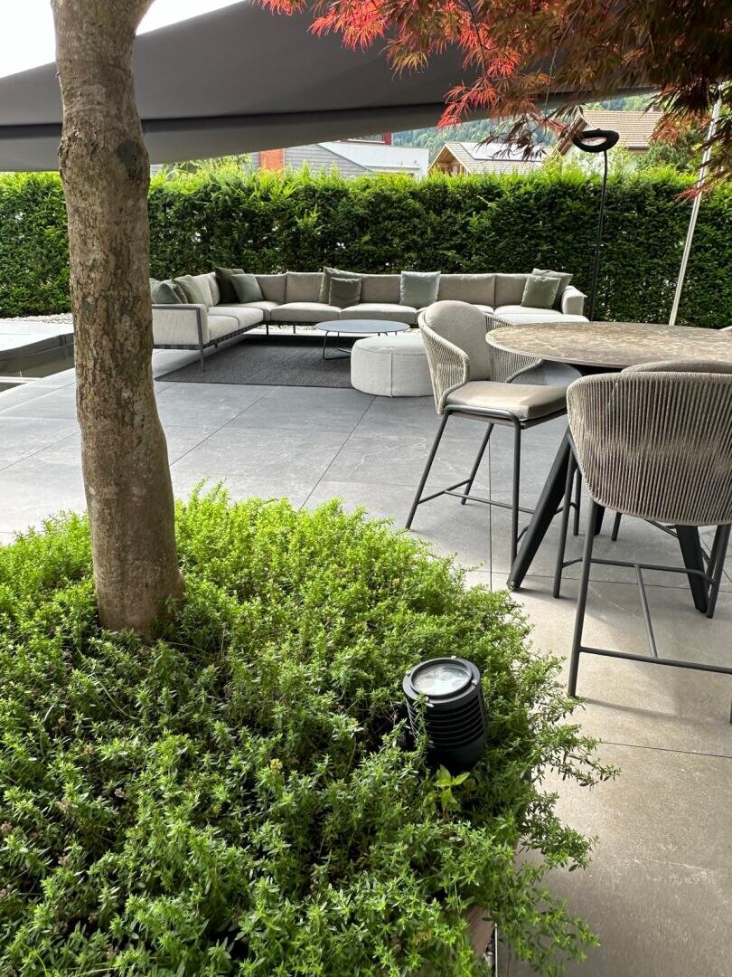 Moderne Außenterrasse mit grauen gepolsterten Sitzmöbeln, einem runden Hocker und einem Esstisch mit Stühlen. Grüne Sträucher und ein Baum stehen im Vordergrund, eine Hecke und ein großer Sonnenschirm spenden Schatten.