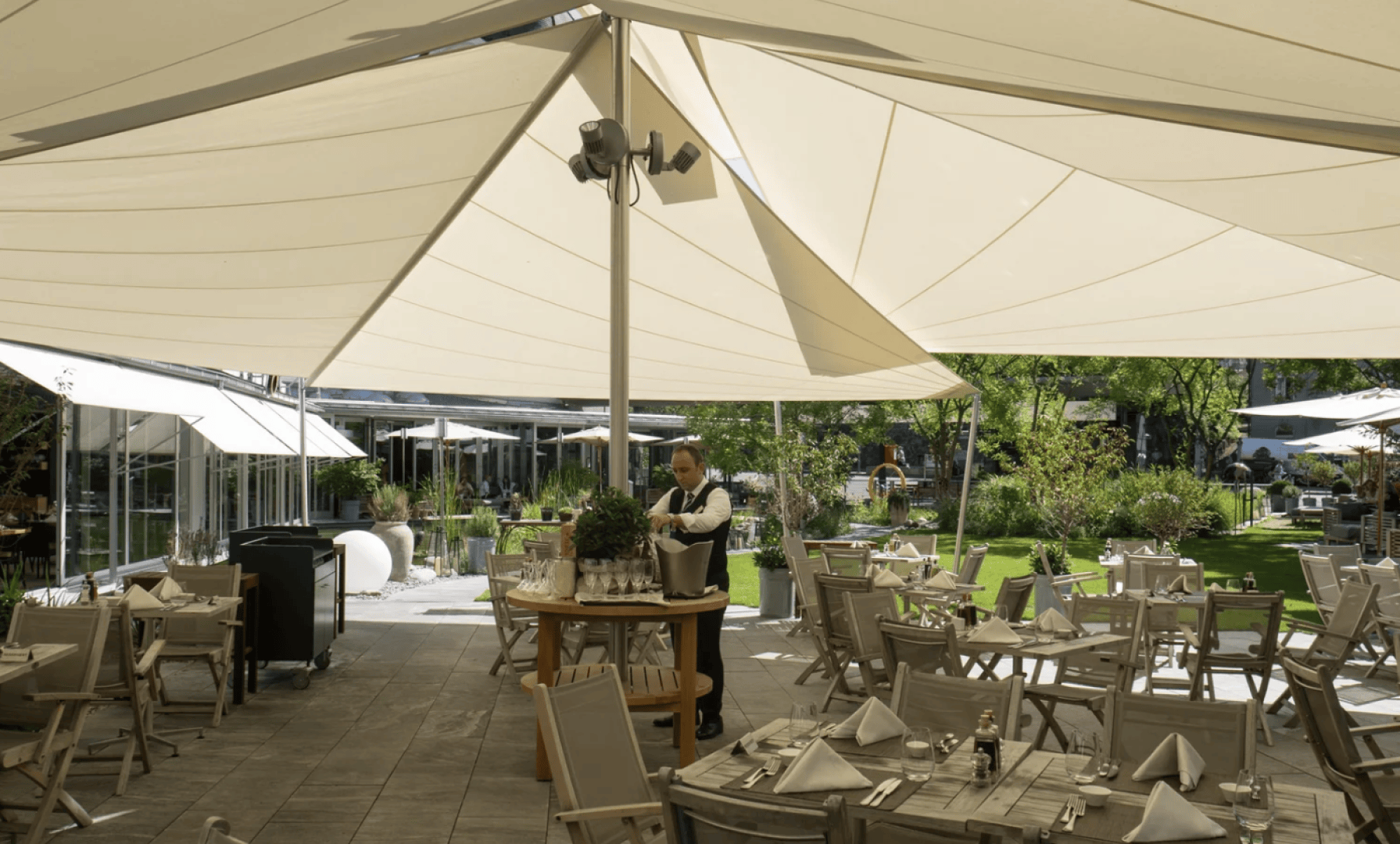 A waiter stands under large cream-colored canopy shades at an outdoor restaurant. Tables and chairs are arranged neatly with folded napkins, and greenery surrounds the patio area.