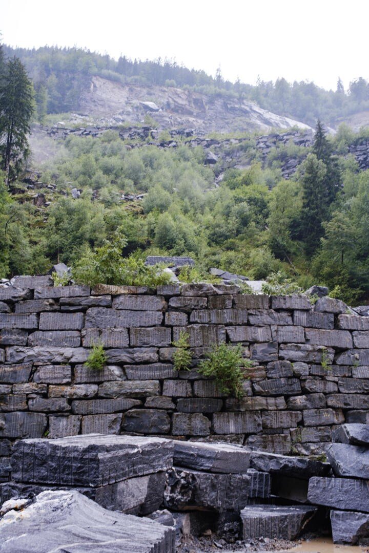 Eine Mauer aus großen, rechteckigen Steinblöcken steht an einem nebligen Tag vor einem grünen, bewaldeten Hang mit verstreuten Felsen und Bäumen.