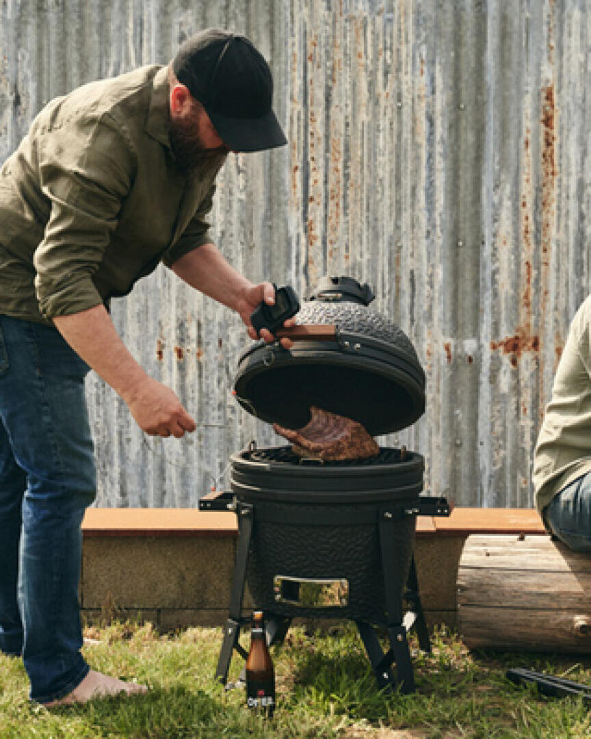 A man opens the lid of a black ceramic barbecue to check if the meat inside is sizzling, outside on the grass, with a corrugated iron wall and a beer bottle nearby. Another person sits partially outside the picture.