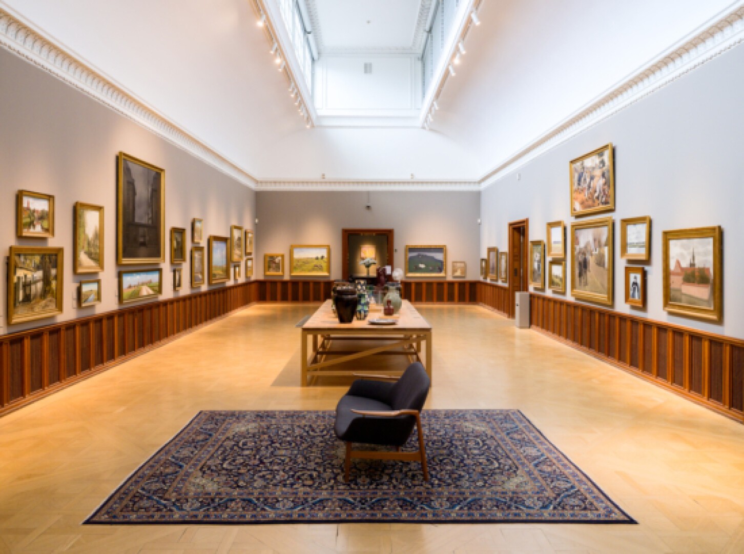 Eine geräumige Kunstgalerie mit gerahmten Gemälden an hellgrauen Wänden, einem Holztisch mit Vasen in der Mitte, einem blau gemusterten Teppich und einem einzelnen Stuhl gegenüber dem Kunstwerk. Natürliches Licht fällt durch ein Deckenfenster ein.