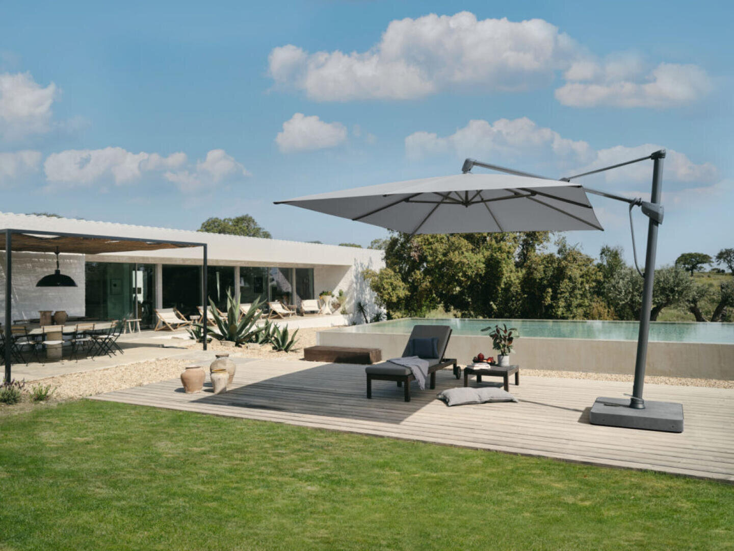 A modern backyard with a pool, lounge chairs, and a large cantilever umbrella on a wooden deck; a covered outdoor dining area and a contemporary house are visible in the background under a partly cloudy sky.