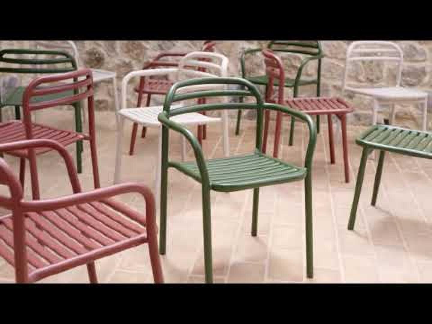 Several metal chairs in green, red and white are randomly arranged on a light-colored brick terrace in front of a stone wall.