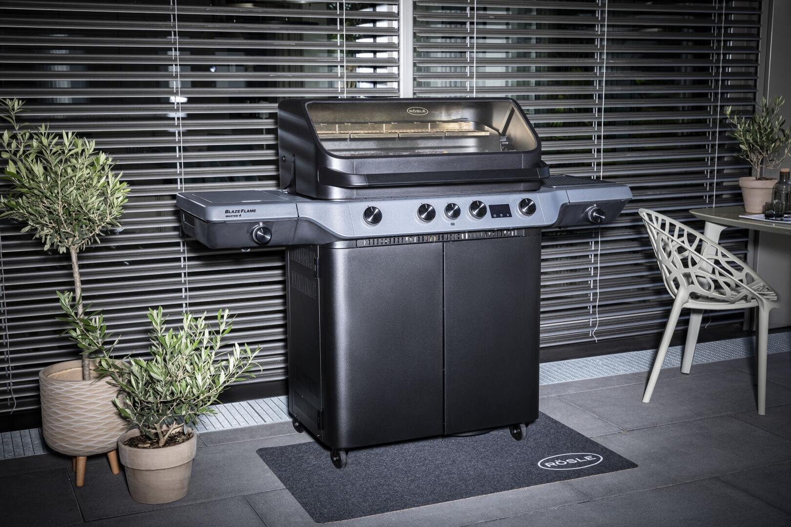 A black outdoor gas barbecue with a closed lid stands on a mat between two potted plants on a tiled terrace. Nearby is a modern chair and a small table, with metal blinds in the background.