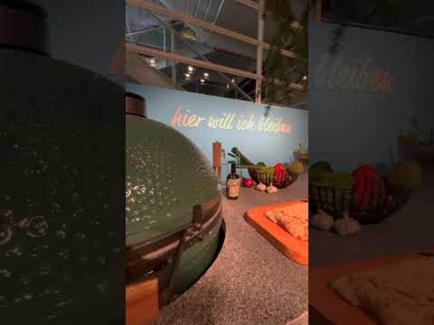 A kitchen counter with a green ceramic grill, chopping boards, fresh produce, garlic bulbs and bottles. On the wall in the background is written in italics: "I want to stay here".