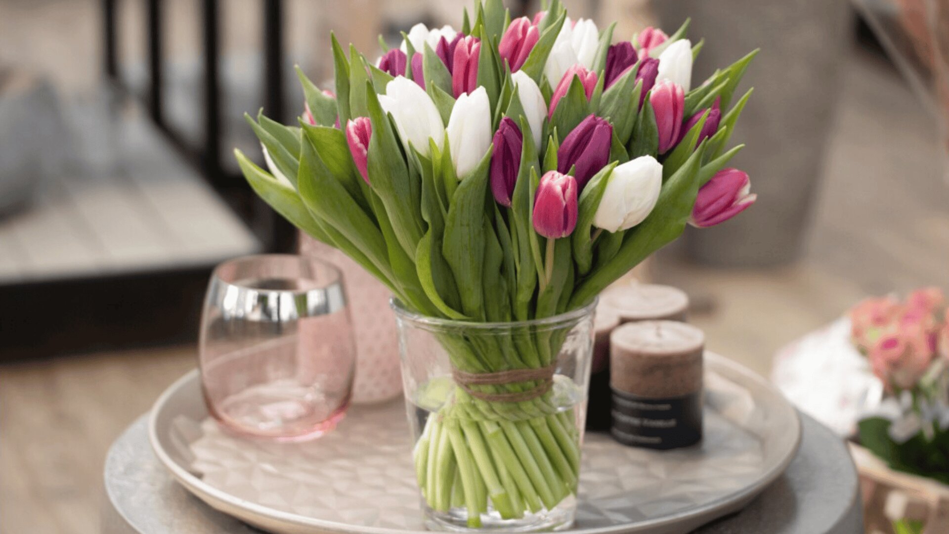 A clear glass vase filled with pink, purple, and white tulips sits on a round tray alongside a pink glass and a brown candle, with flowers blurred in the background.