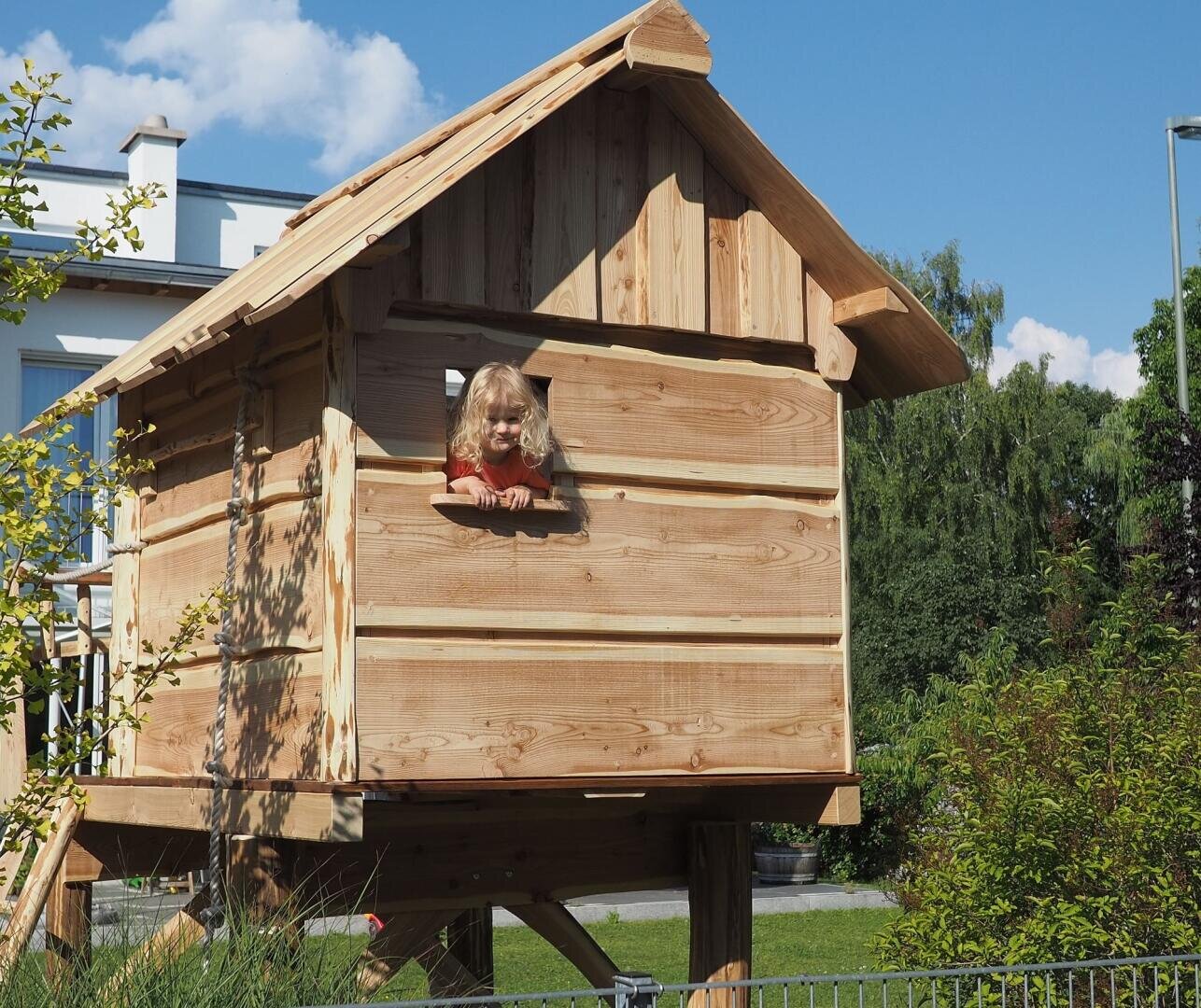 Ein kleines Kind mit blondem Haar lehnt sich lächelnd aus dem Fenster eines hölzernen Baumhauses auf Stelzen. Das Baumhaus steht in einem grünen Garten mit Sträuchern, und im Hintergrund ist ein modernes Haus zu sehen.