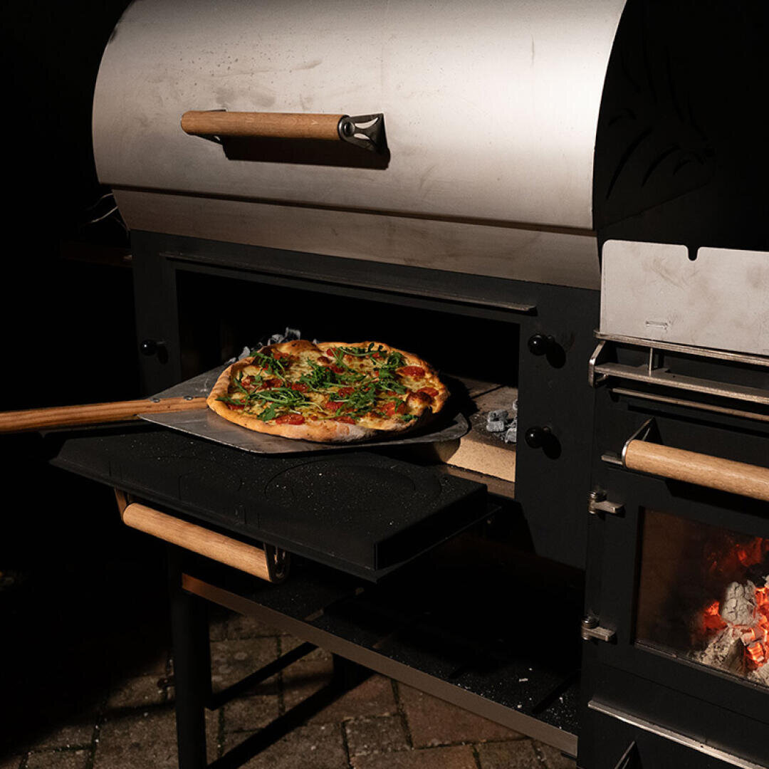 Eine frisch gebackene Pizza auf einer hölzernen Pizzaschaufel wird aus einem metallenen Pizzaofen im Freien geholt, in dem ein Feuer in einem Seitenfach brennt.