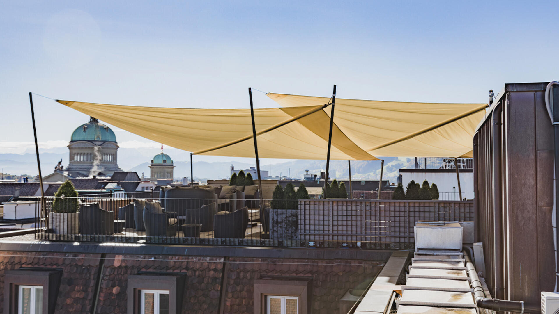 Dachterrasse mit Sitzgelegenheiten und großen, gelben Sonnensegeln, mit Blick auf die Gebäude und Kuppeln der Stadt unter einem klaren blauen Himmel. Im Hintergrund sind die Berge in der Ferne zu sehen.
