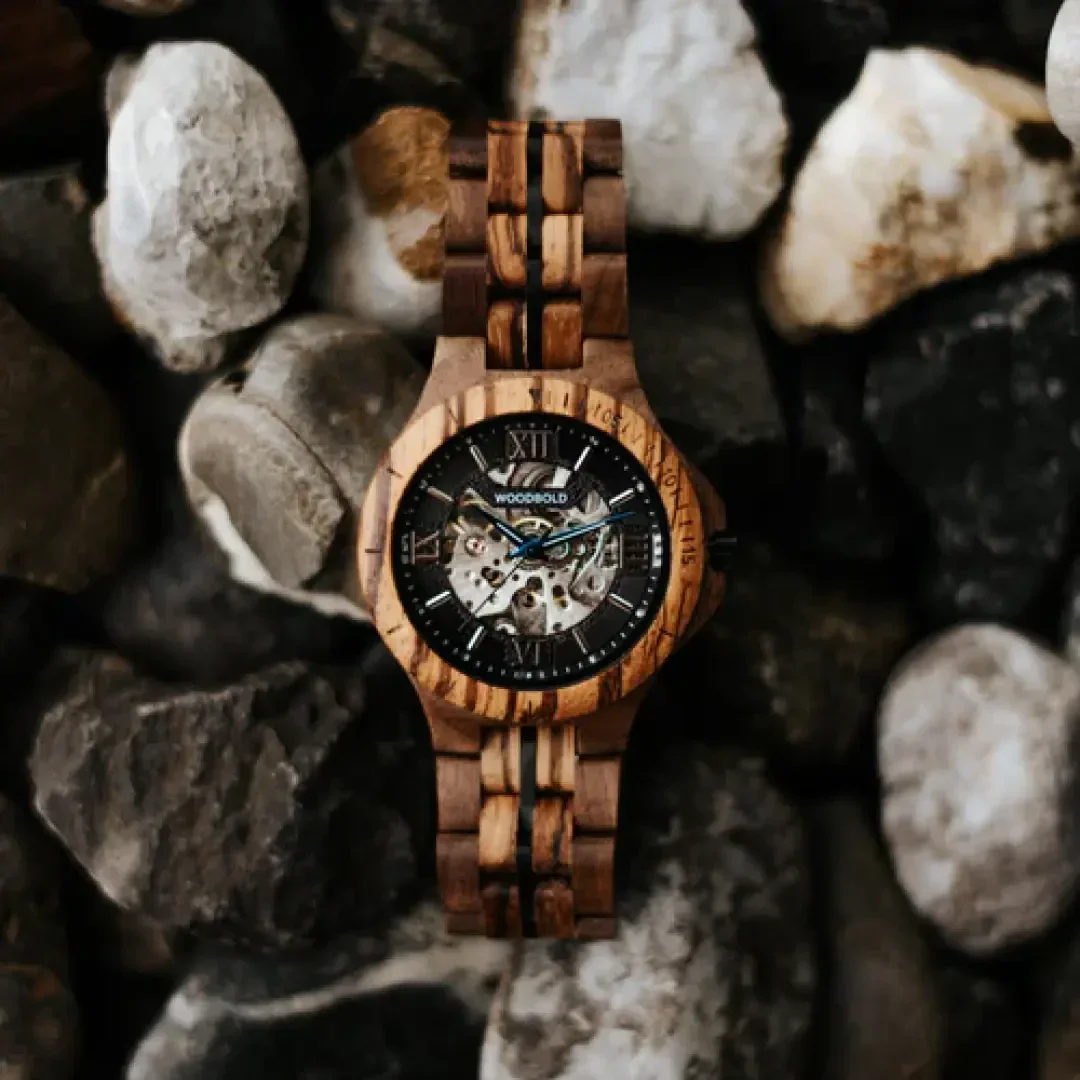 A wooden wristwatch with a dark brown and light brown strap and a black skeleton dial with cogwheels lies on a bed of different colored smooth stones.