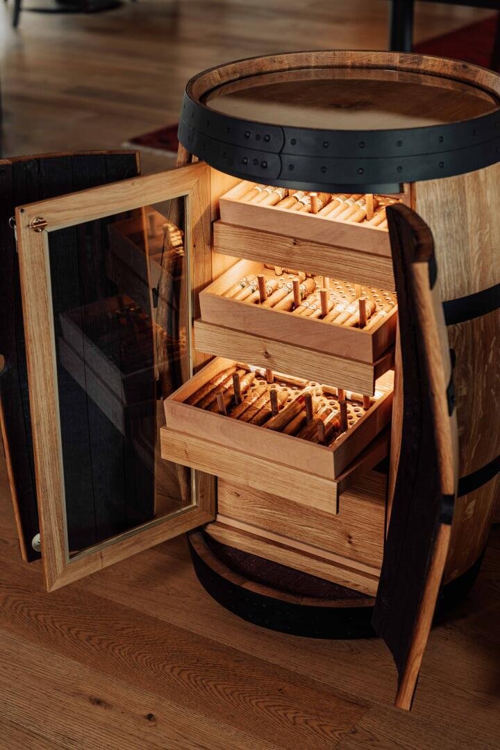 A humidor made of wooden barrels with a glass door is open and provides a view of several illuminated wooden drawers, neatly filled with cigars and placed on a wooden floor.