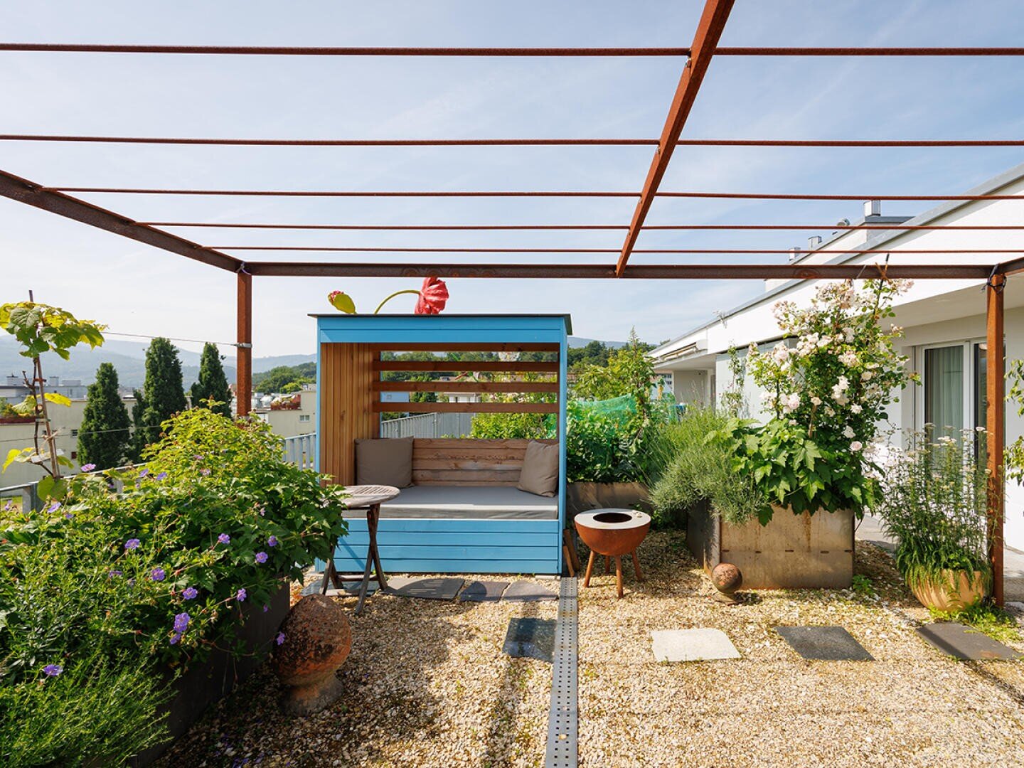 Eine Dachterrasse mit einer blauen hölzernen Sitzecke, einem kleinen Tisch, Topfpflanzen und einem Pergola-Gestell aus Metall. Der Bereich ist mit Grünpflanzen dekoriert und hat einen Kiesboden unter einem klaren, sonnigen Himmel.