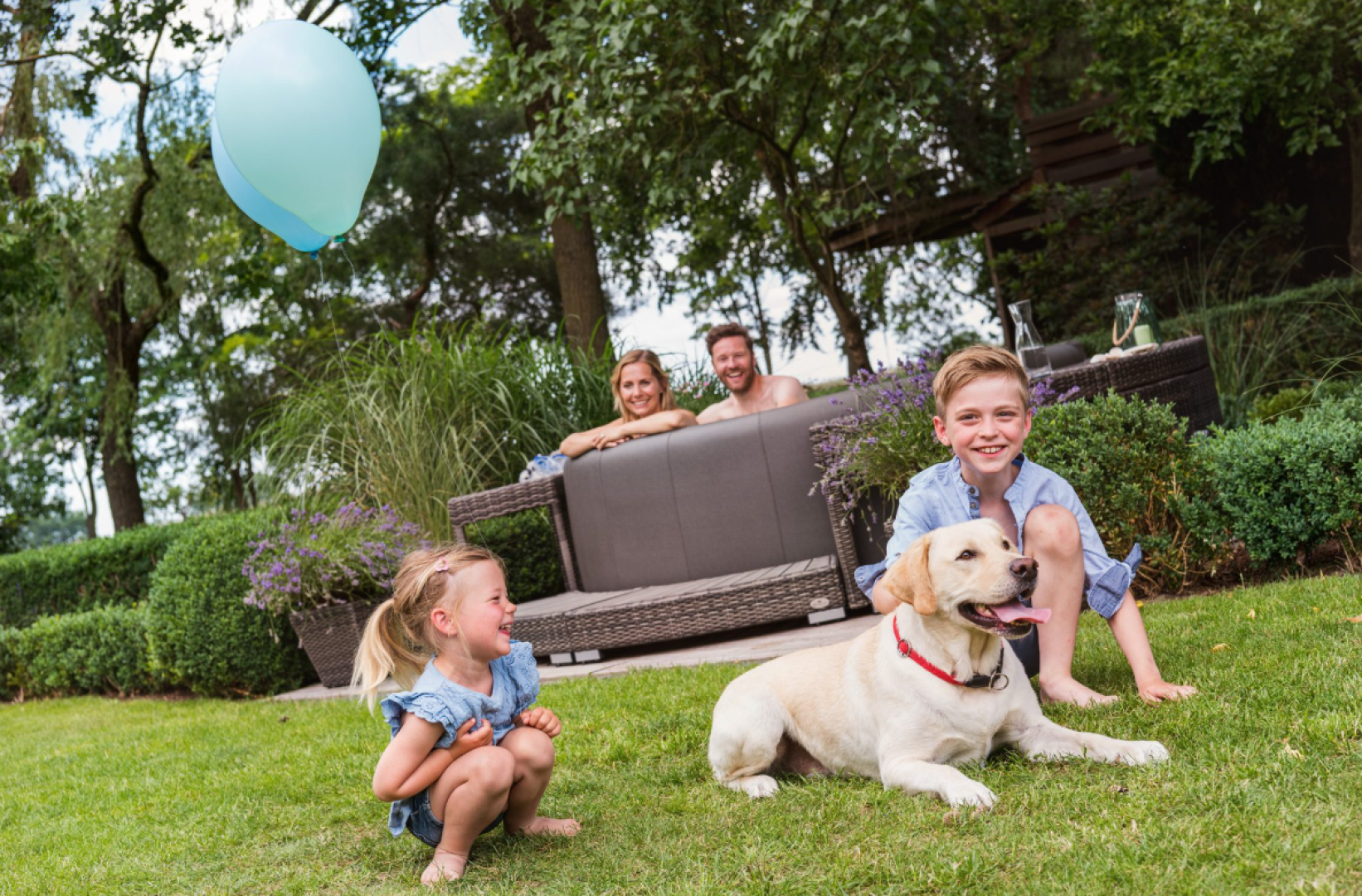 Zwei Kinder und ein gelber Labradorhund spielen im Gras, während sich zwei Erwachsene auf einem Gartensofa im Hintergrund entspannen; ein blauer Luftballon schwebt an einem sonnigen Tag über ihnen.