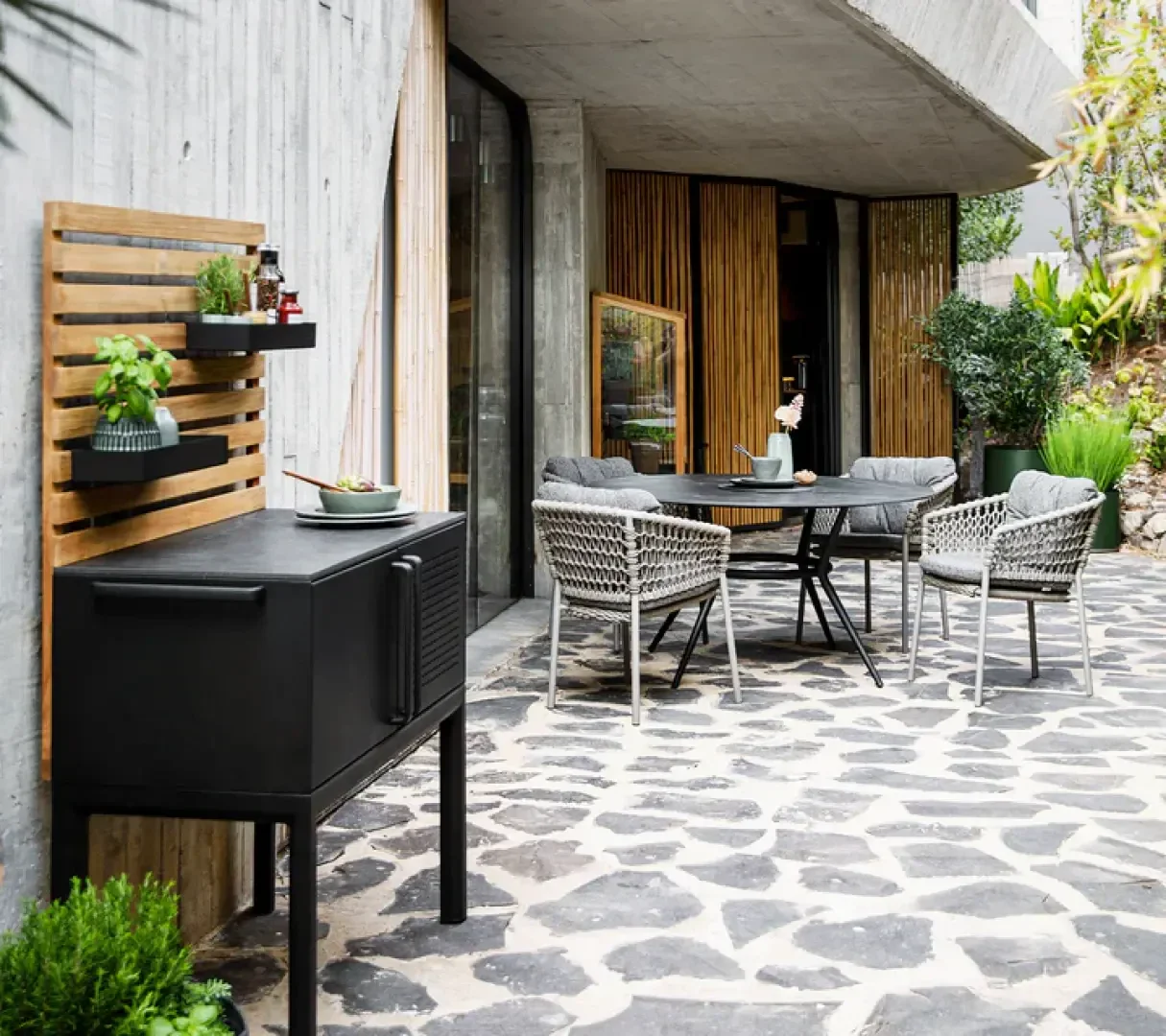 Auf einer modernen Terrasse steht ein runder Tisch mit vier geflochtenen Stühlen, Plüschkissen und Topfpflanzen. In der Nähe steht ein schwarzer Außengrill mit Kräutern und Dekoration an einer Holzlattenwand. Der Boden des Patios besteht aus unregelmäßigen Steinfliesen.