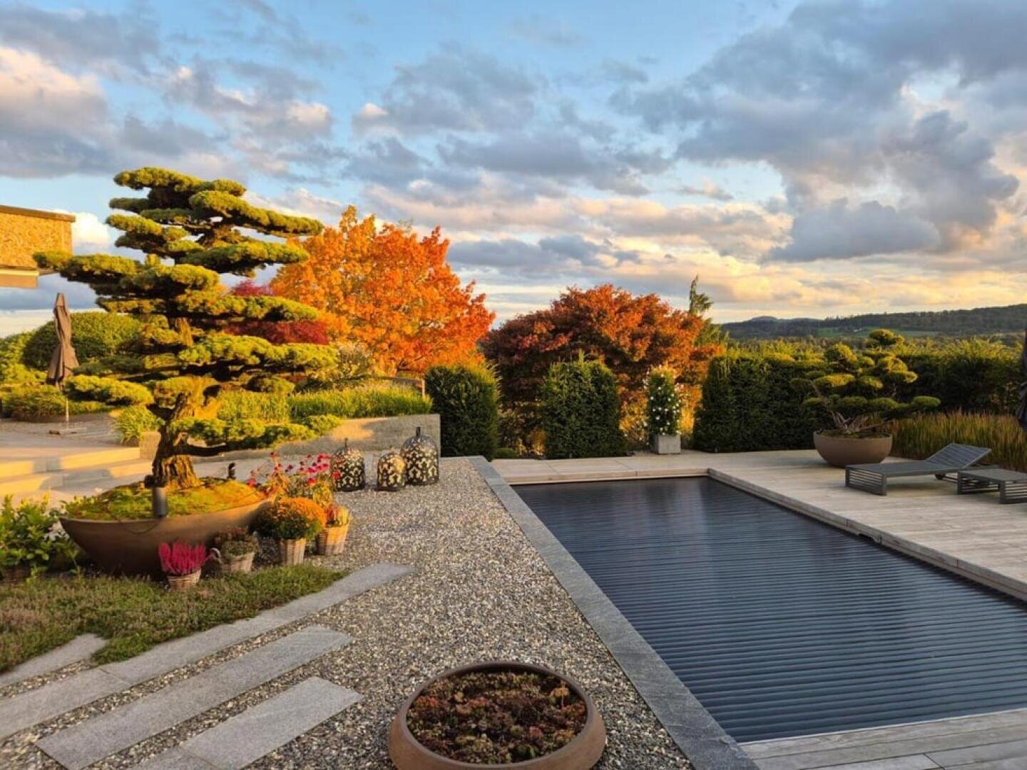 Im Garten gibt es einen überdachten Pool, gepflegte Bäume in großen Töpfen, farbenfrohes Herbstlaub, Steintreppen, Topfblumen und Liegestühle, und das alles unter einem teilweise bewölkten Himmel mit warmem Sonnenlicht.
