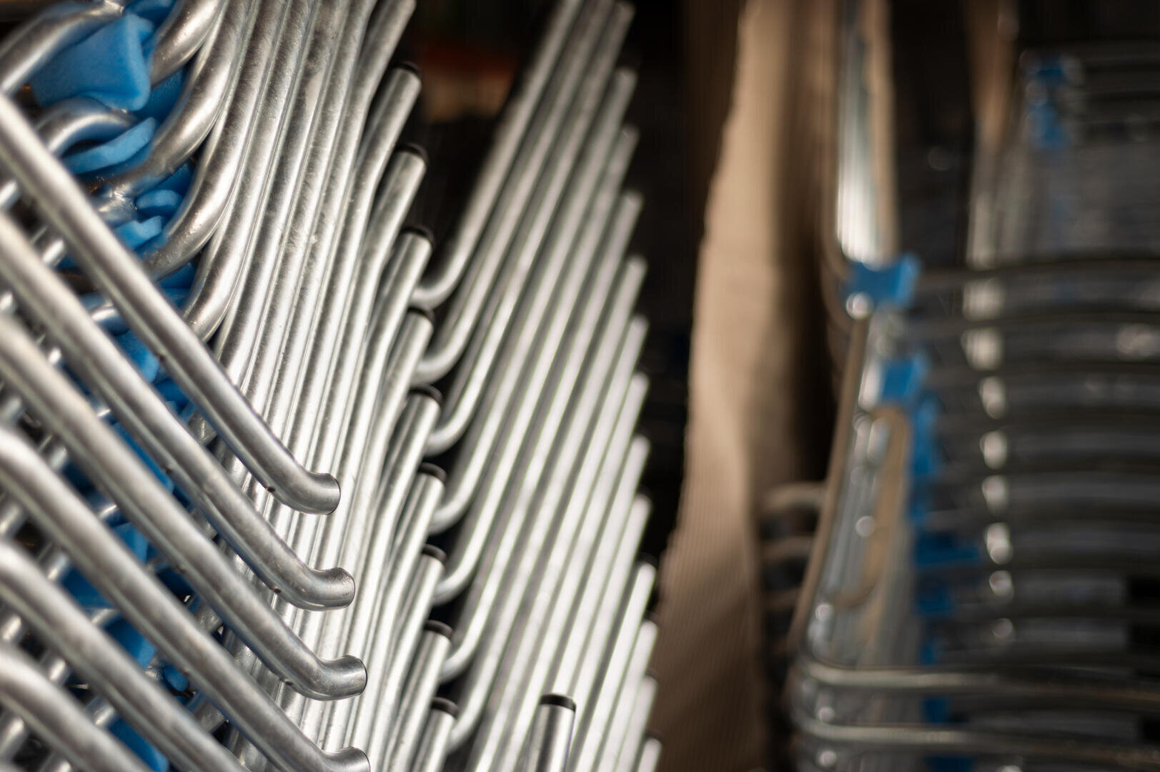 Stapel von Metallstühlen sind dicht aneinandergereiht, so dass ein Muster aus silbern glänzenden Beinen und blauen Sitzen entsteht. Das Foto ist schräg aufgenommen, wodurch die Wiederholung und die Linien der Stuhlrahmen betont werden.