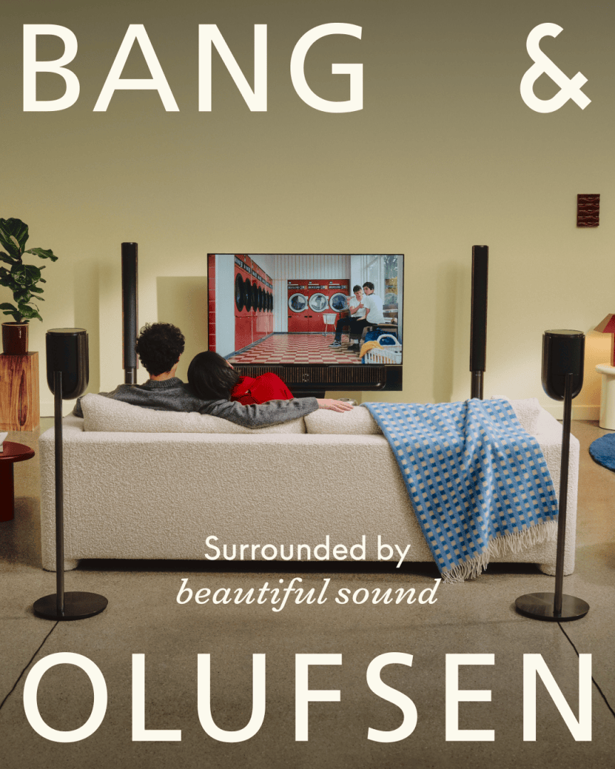 Ein Paar sitzt auf einer weißen Couch und sieht fern, während um sie herum Surround-Sound-Lautsprecher aufgestellt sind. BANG & OLUFSEN und Surrounded by beautiful sound sind in großer Schrift auf dem Bild zu sehen.