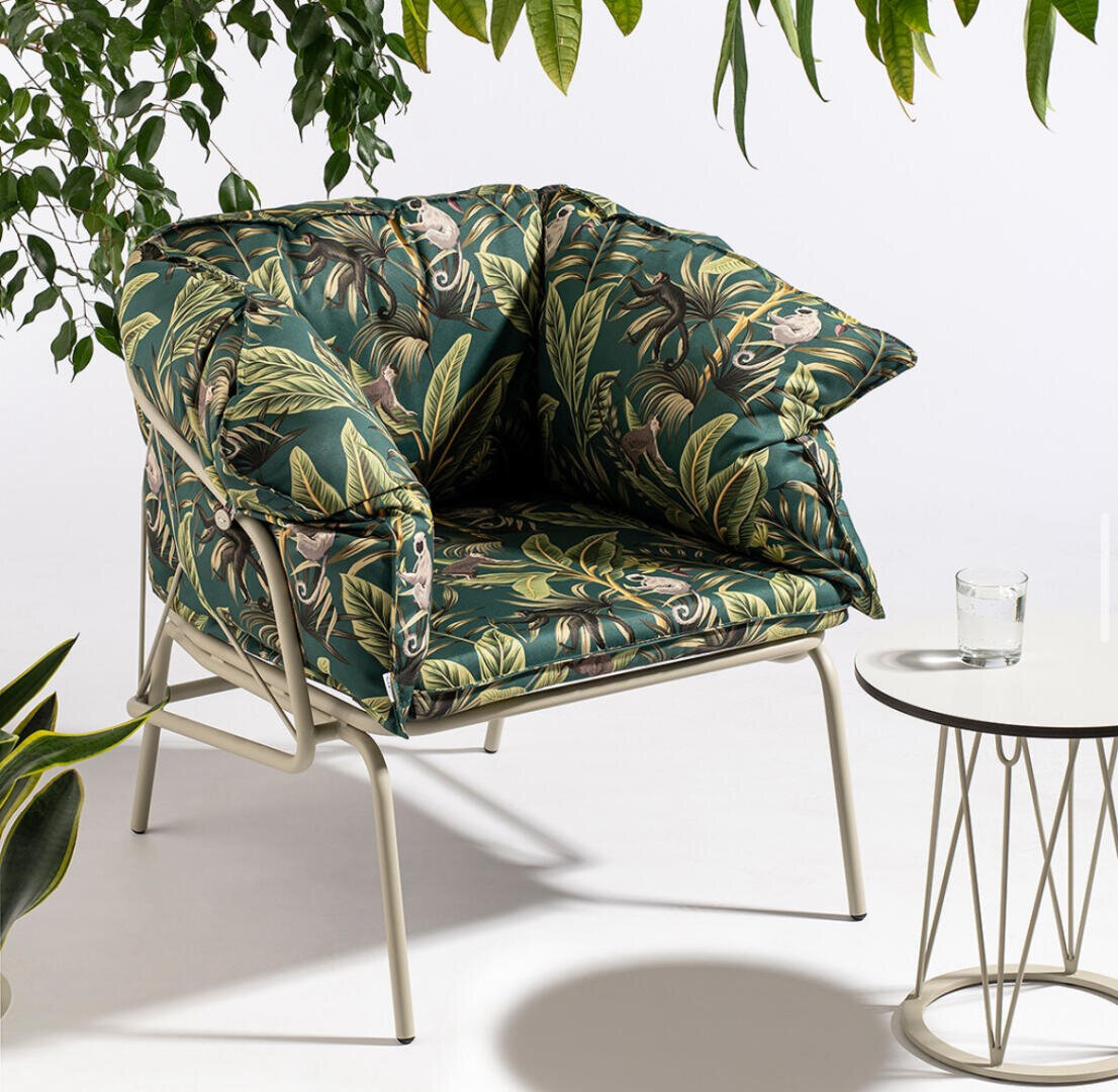 Ein gepolsterter Sessel mit einem grünen tropischen Blattmuster steht neben einem runden weißen Tisch mit dünnen Metallbeinen. Auf dem Tisch steht ein Glas Wasser, und über und um den Sessel hängen Blattpflanzen.