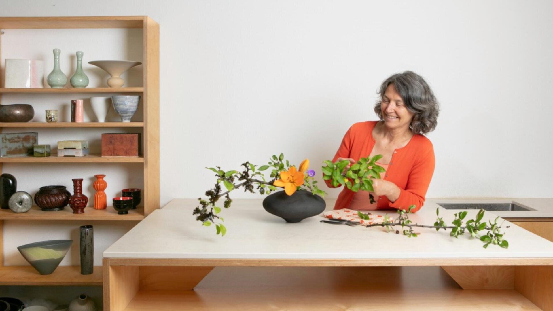 Eine Frau mit grauem Haar und einer orangefarbenen Strickjacke arrangiert lächelnd einen Blumenstrauß in einer schwarzen Vase auf einem weißen Tisch. Hinter ihr steht ein Holzregal mit dekorativen Töpferwaren und Vasen.