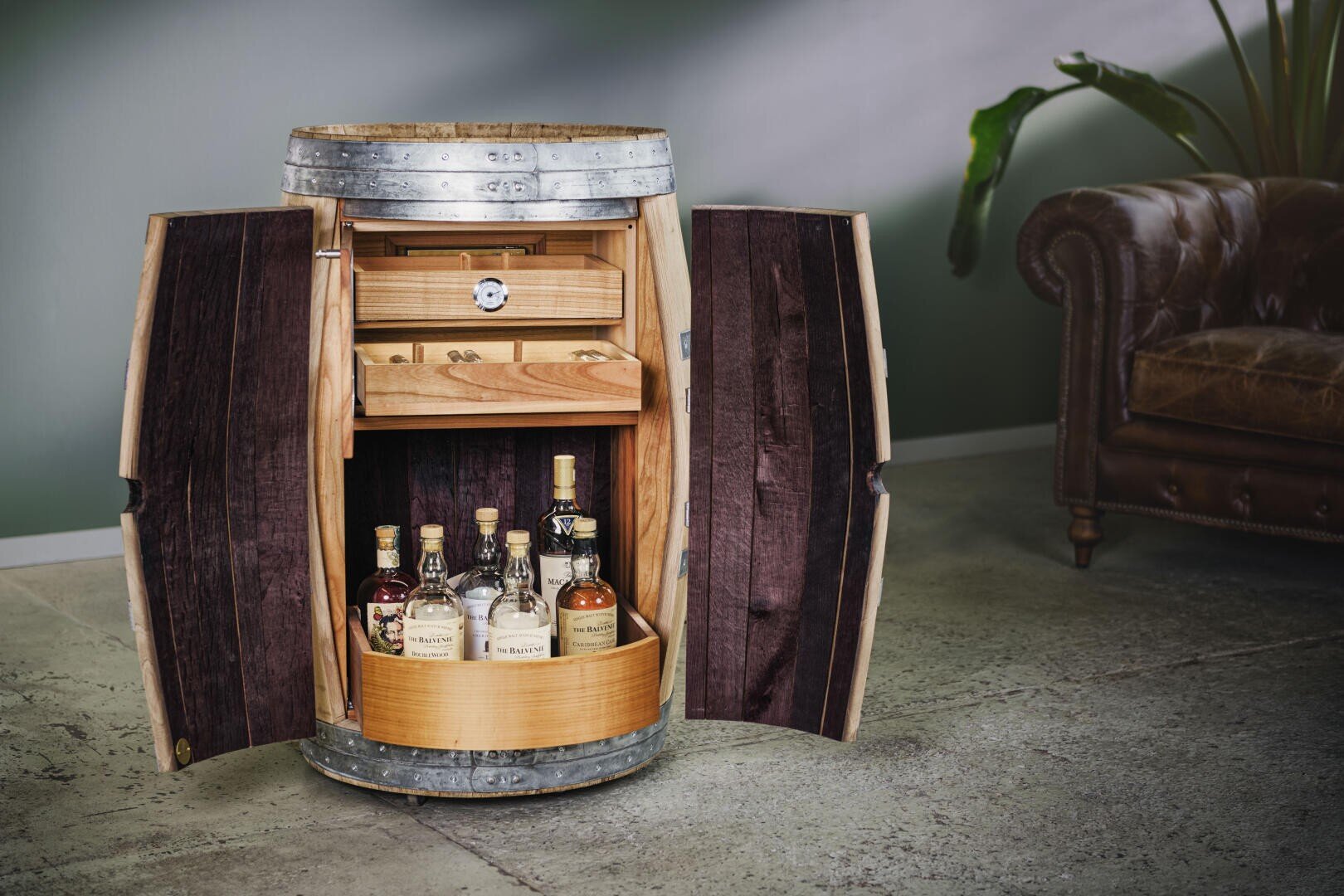 A wooden barrel that has been converted into a cupboard with open doors, with bottles of schnapps on the bottom shelf and a drawer above, next to a brown leather sofa and a green plant in a modern room.