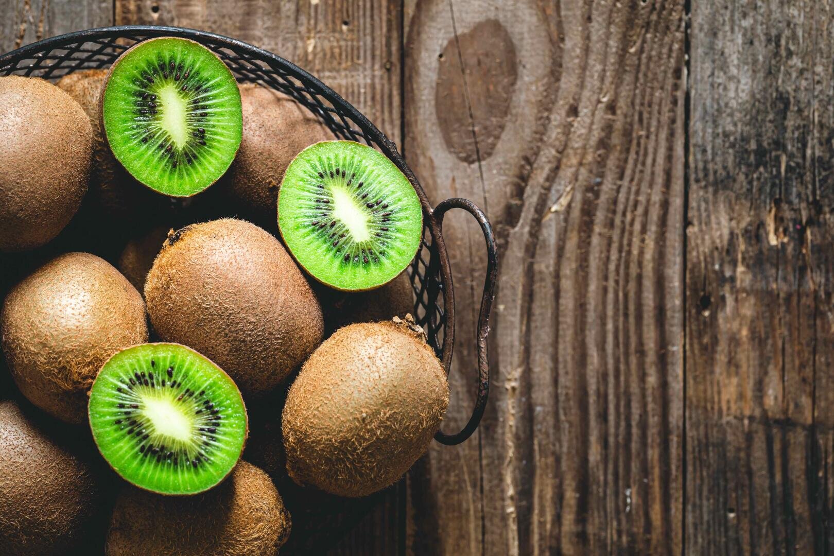 A basket with whole kiwis and two halved kiwis, showing their bright green insides, stands on a rustic wooden surface.