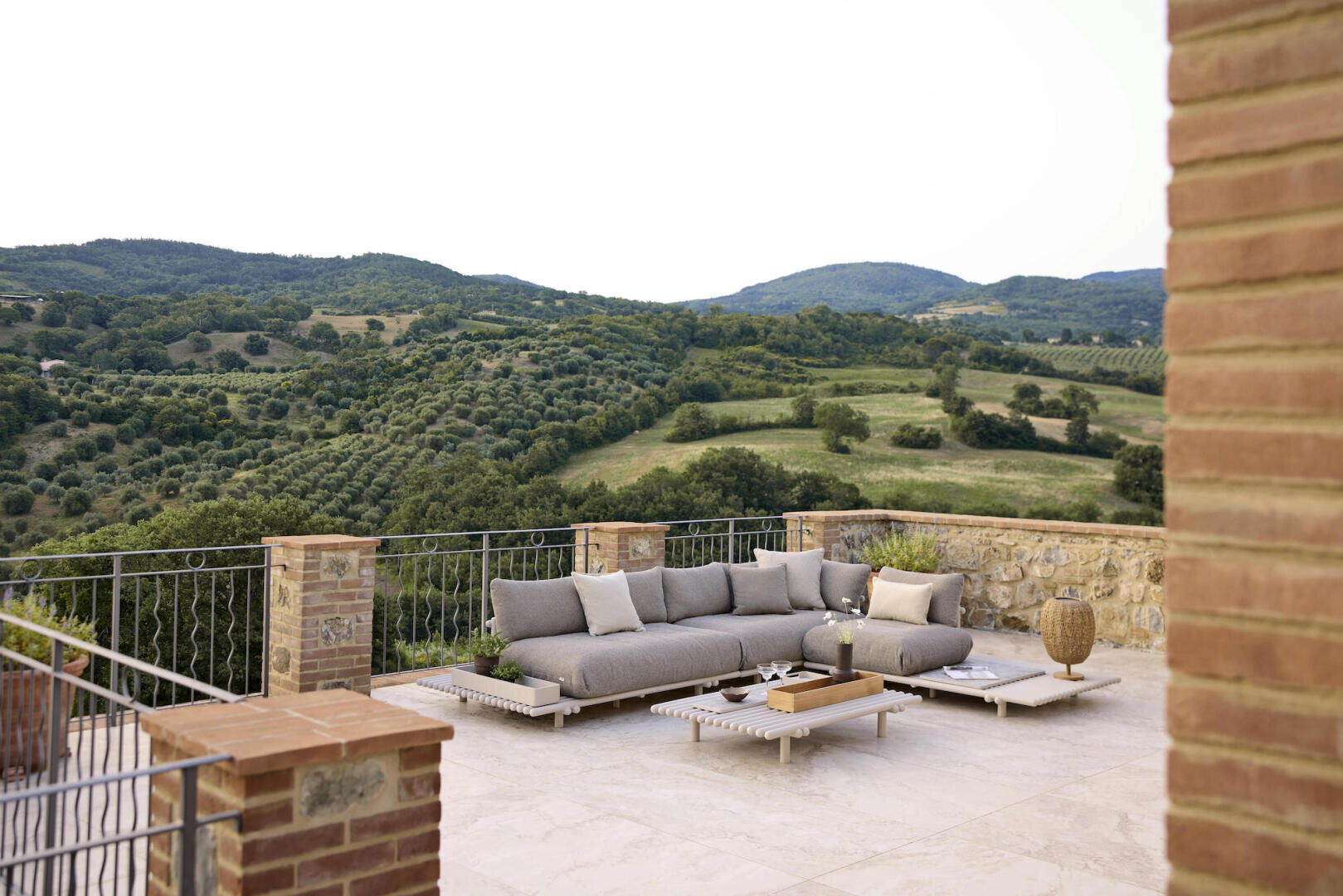 Eine moderne Terrasse mit grauen Sofas und Kissen, die auf einer Steinterrasse mit Metallgeländer steht, bietet einen Blick auf grüne Hügel und eine malerische Landschaft unter freiem Himmel.