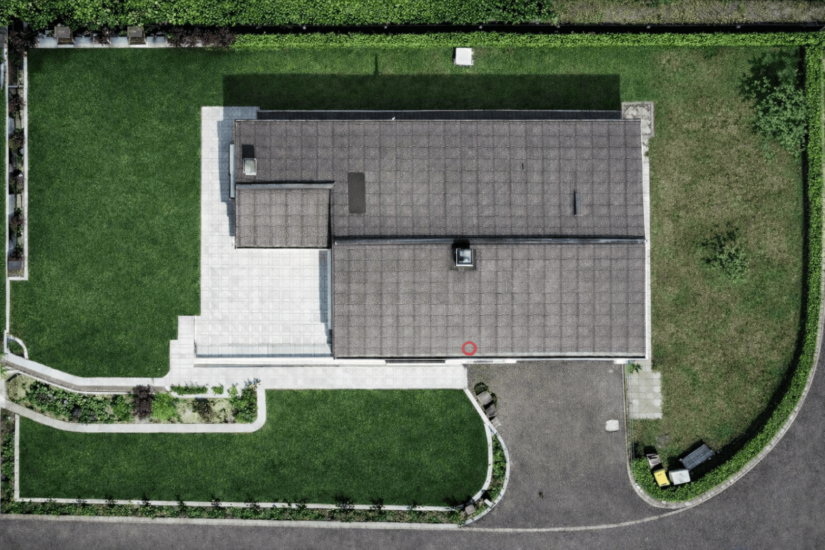 Luftaufnahme eines modernen Hauses mit grauem Dach, umgeben von einer grünen Rasenfläche, einer Auffahrt und Hecken. Ein kleiner roter Kreis markiert eine Stelle auf dem Dach in der Nähe der unteren Mitte.
