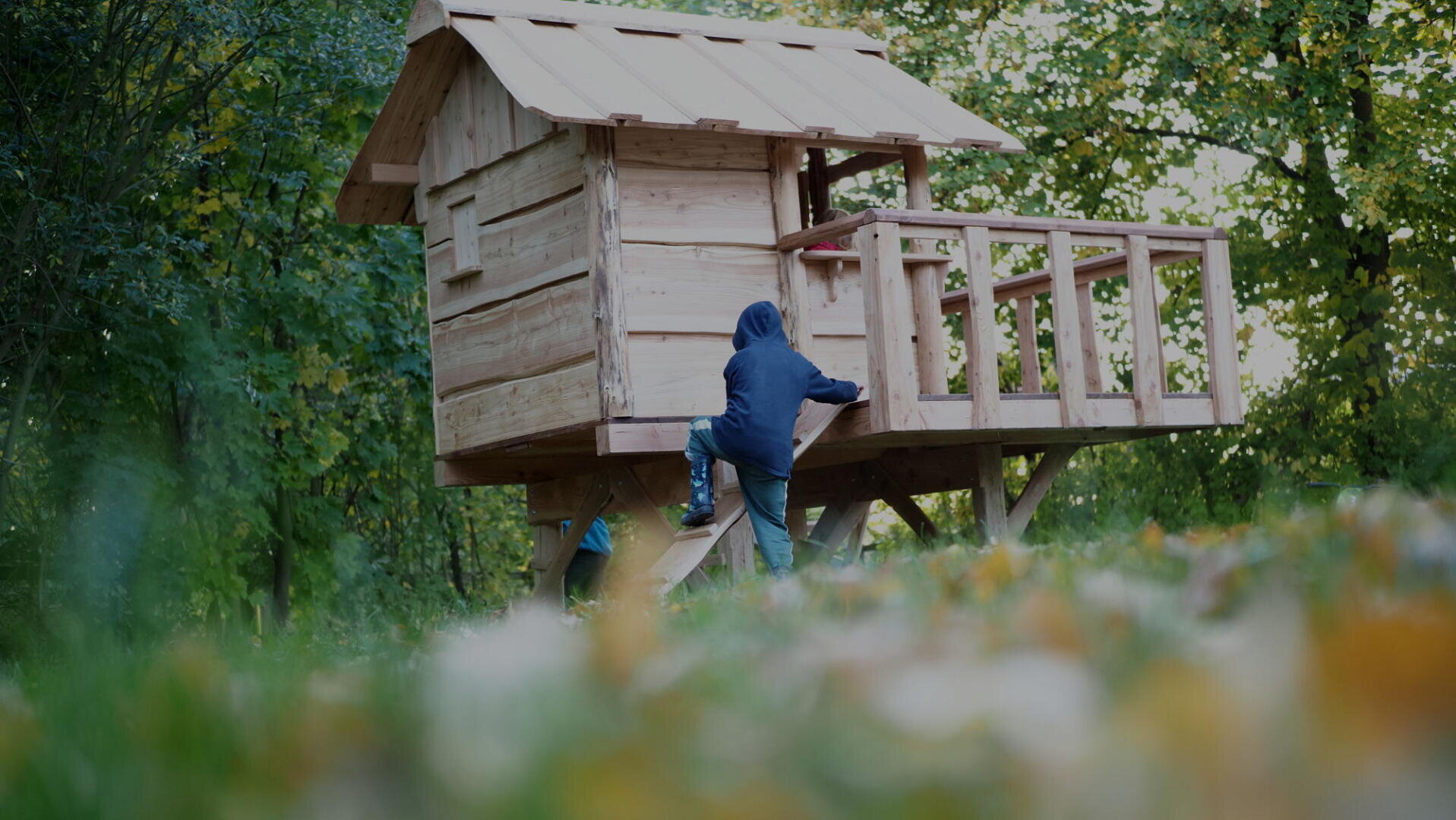 A child in a blue hoodie climbs the ladder of a wooden treehouse surrounded by green trees and grass.