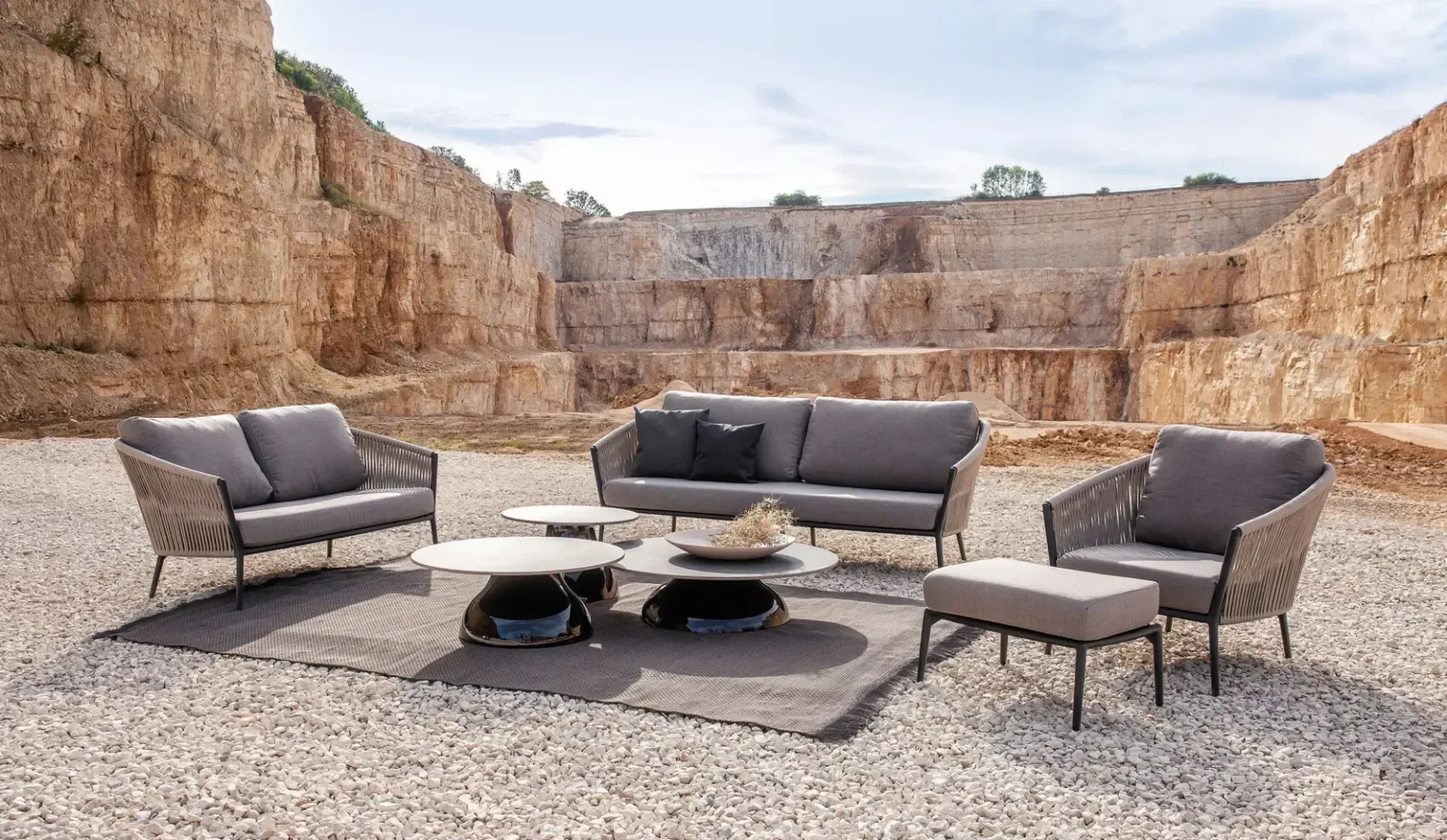 A modern outdoor furniture set with a sofa, two armchairs, an ottoman, and two round coffee tables is arranged on a rug in a rocky, open quarry under a partly cloudy sky.
