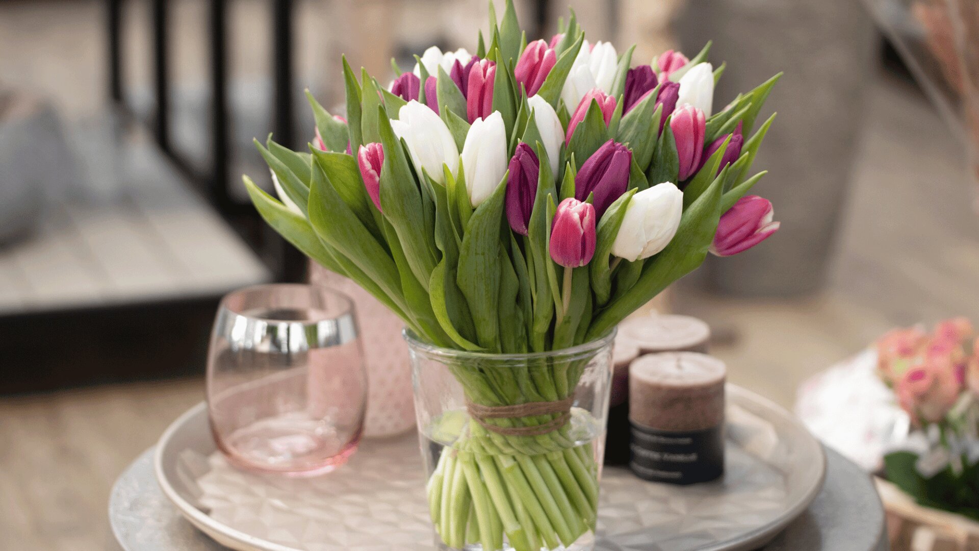 Eine Klarglasvase enthält einen Strauß weißer und violetter Tulpen mit grünen Blättern, der auf einem Tablett neben einem rosa Glas und zwei braunen Kerzen steht. Der Hintergrund ist sanft verschwommen.