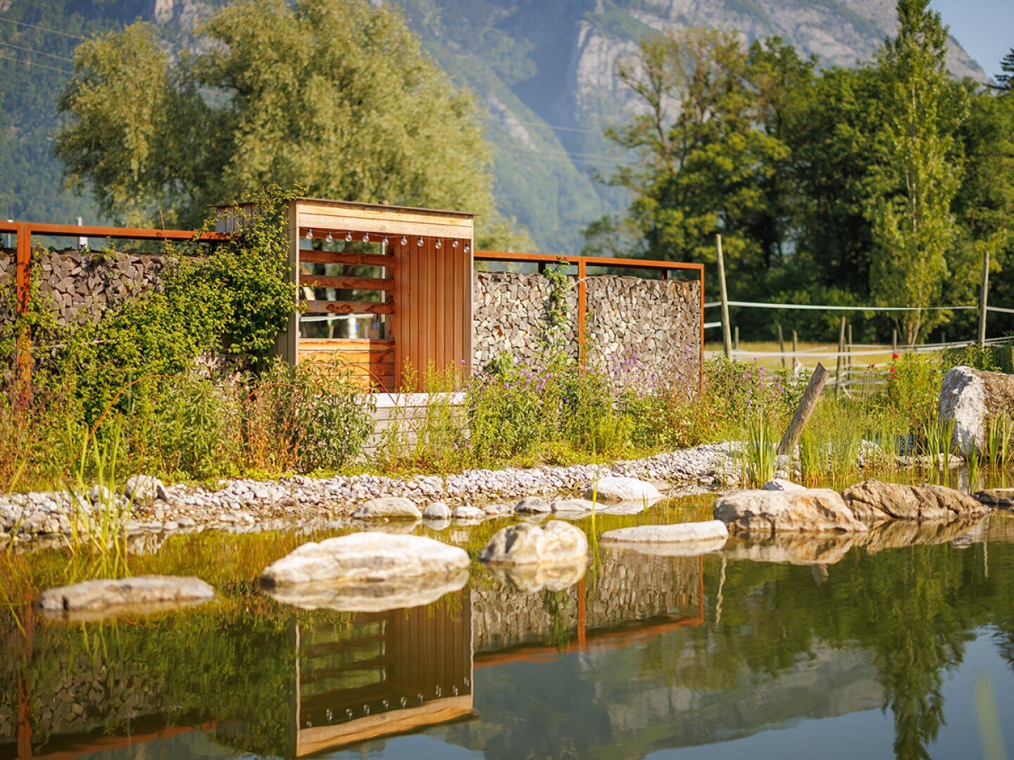 Ein natürlicher Teich mit großen Steinen am Rand und klarem Wasser, in dem sich ein Holztor und ein mit Steinen gefüllter Zaun spiegeln, umgeben von üppigem Grün und fernen Bergen.