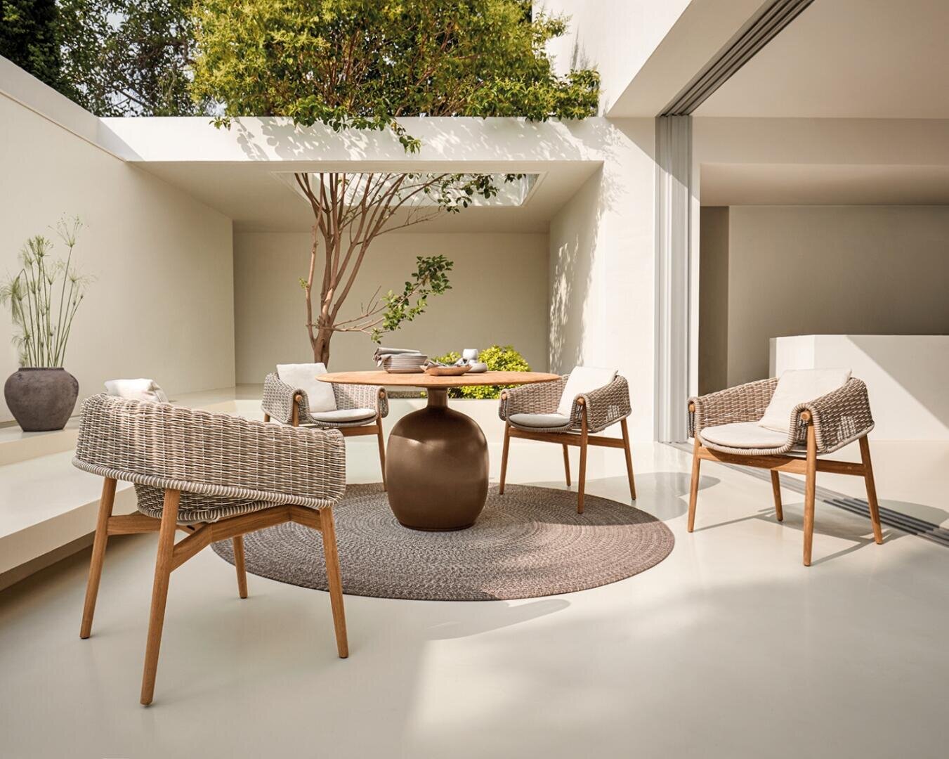Eine moderne Außenterrasse mit einem runden Tisch, vier Korbstühlen und einem runden Teppich, umgeben von weißen Wänden, Pflanzen und einem Baum, die eine helle und ruhige Atmosphäre schaffen.
