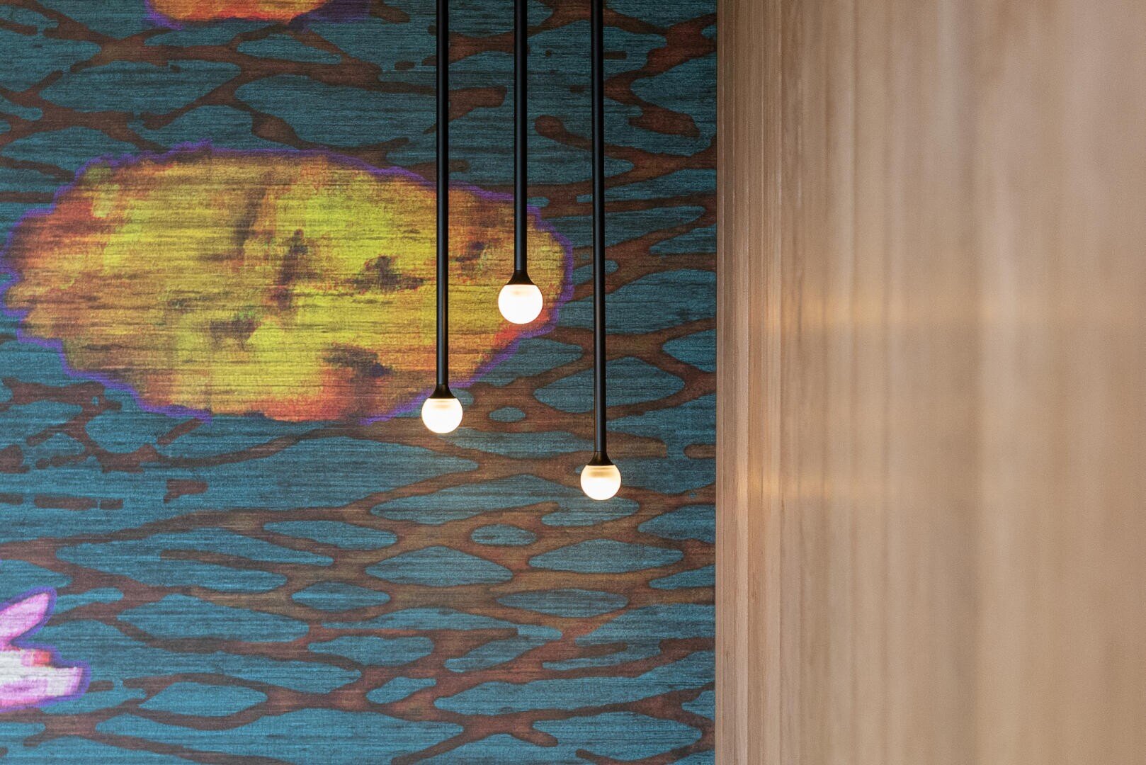 Three hanging light bulbs are suspended in front of a colorful, abstract wall with blue, yellow and brown patterns. A plain wooden surface can be seen on the right-hand side of the picture.