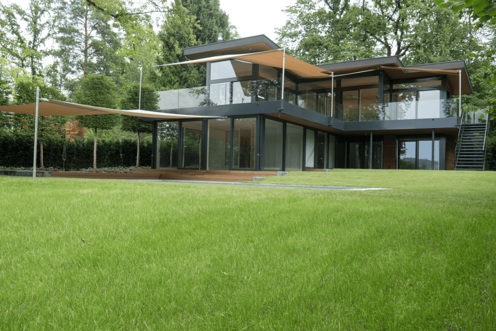 Ein modernes zweistöckiges Haus mit großen Glasfenstern, Flachdächern und hölzernen Akzenten ist von üppigem grünen Gras und Bäumen umgeben. Das Haus verfügt über Außenterrassen und mehrere Überhänge, die Schatten spenden.