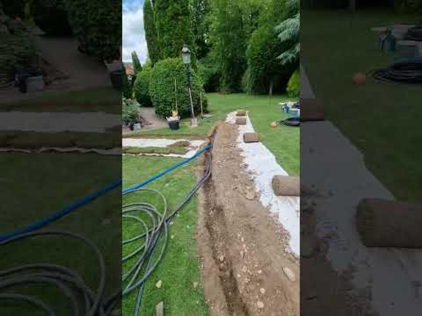 Ein Garten, in dem ein langer Graben durch das Gras ausgehoben wurde, in dessen Nähe Rollen von abgetragenem Torf liegen, Gartengeräte und Schläuche verstreut sind und Bäume und Sträucher im Hintergrund zu sehen sind.