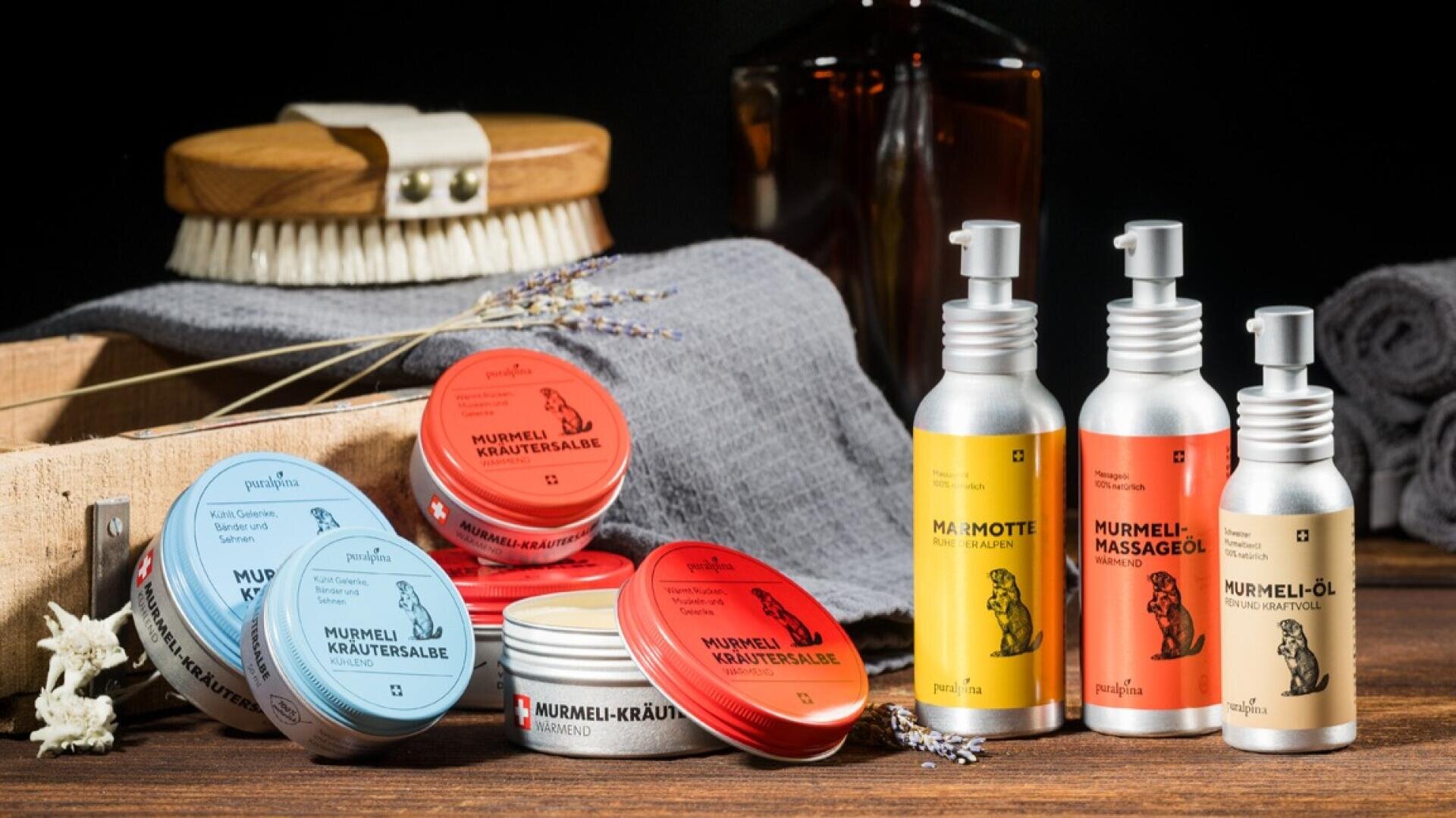 Eine Auslage mit verschiedenen Kräutersalben und Massageölen in bunten Dosen und Flaschen mit Tierdarstellungen, angeordnet auf einer hölzernen Fläche mit einem Pinsel, einem grauen Tuch und getrocknetem Lavendel im Hintergrund.