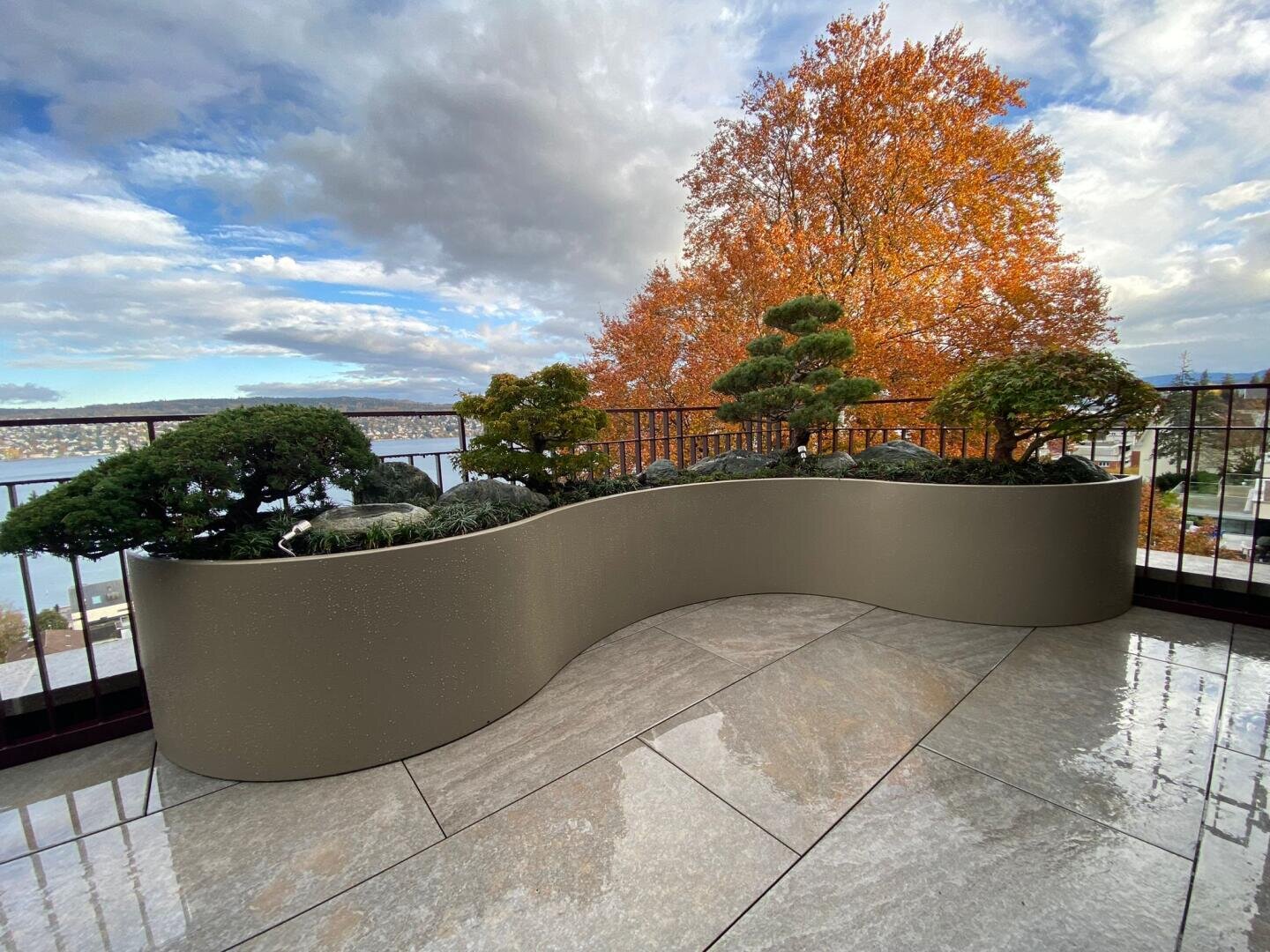 Geschwungenes Pflanzgefäß mit kleinen, gepflegten Bonsai-Bäumen auf einem gefliesten Balkon; im Hintergrund sind ein orangefarbener Baum und ein See mit bewölktem Himmel zu sehen.