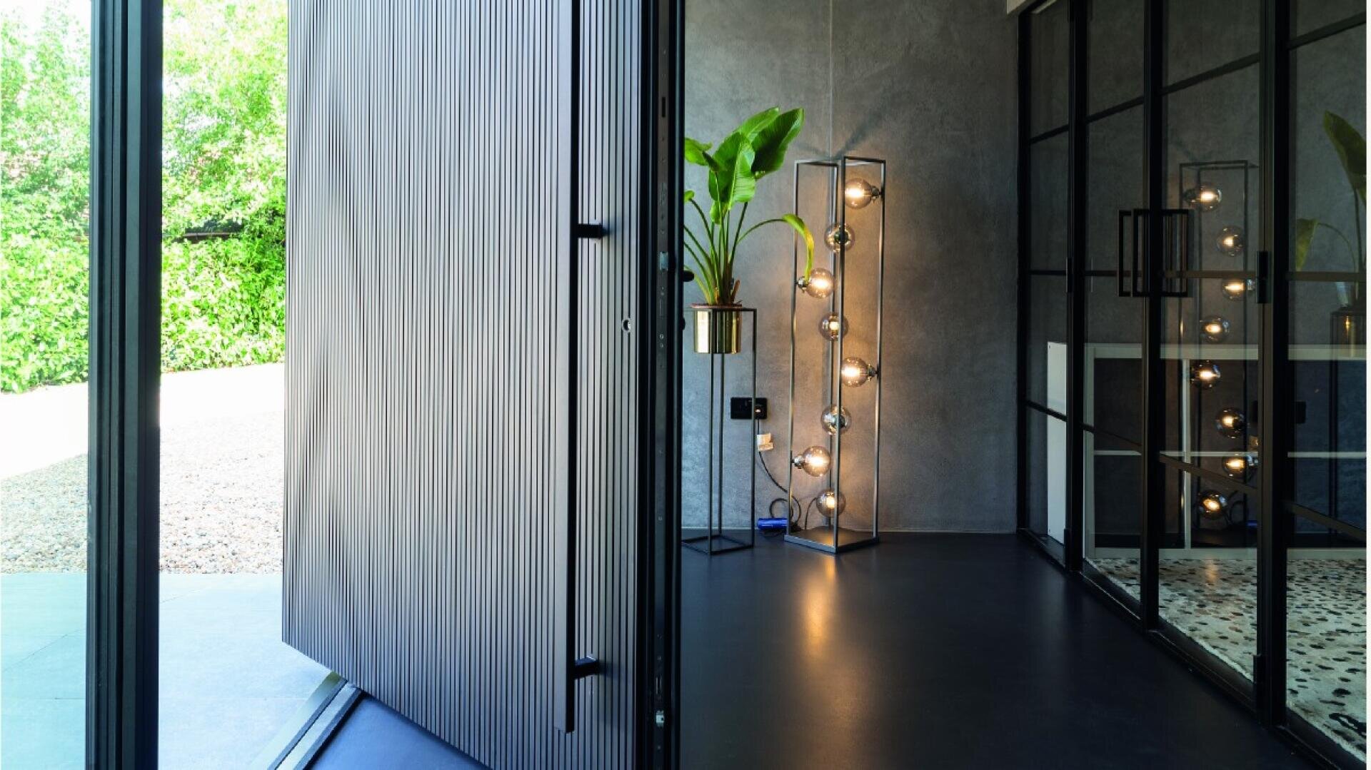 A modern interior with a large swing door open to the outside, black shelves, a potted plant, a floor lamp with exposed light bulbs and glass partitions with black frames. Natural light floods in from outside.