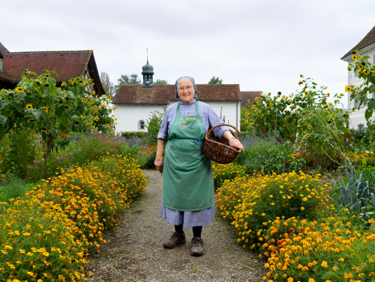 Eine ältere Schwester mit grüner Schürze und einem gefüllten Korb im Arm steht auf einem mit leuchtend gelben Blumen gesäumter Kiesweg, der durch einen üppigen Garten zu einem weissen Gebäude mit rotem Ziegeldach und kleiner Kuppel führt, das von Sonnenblumen und Grünpflanzen umgeben ist.