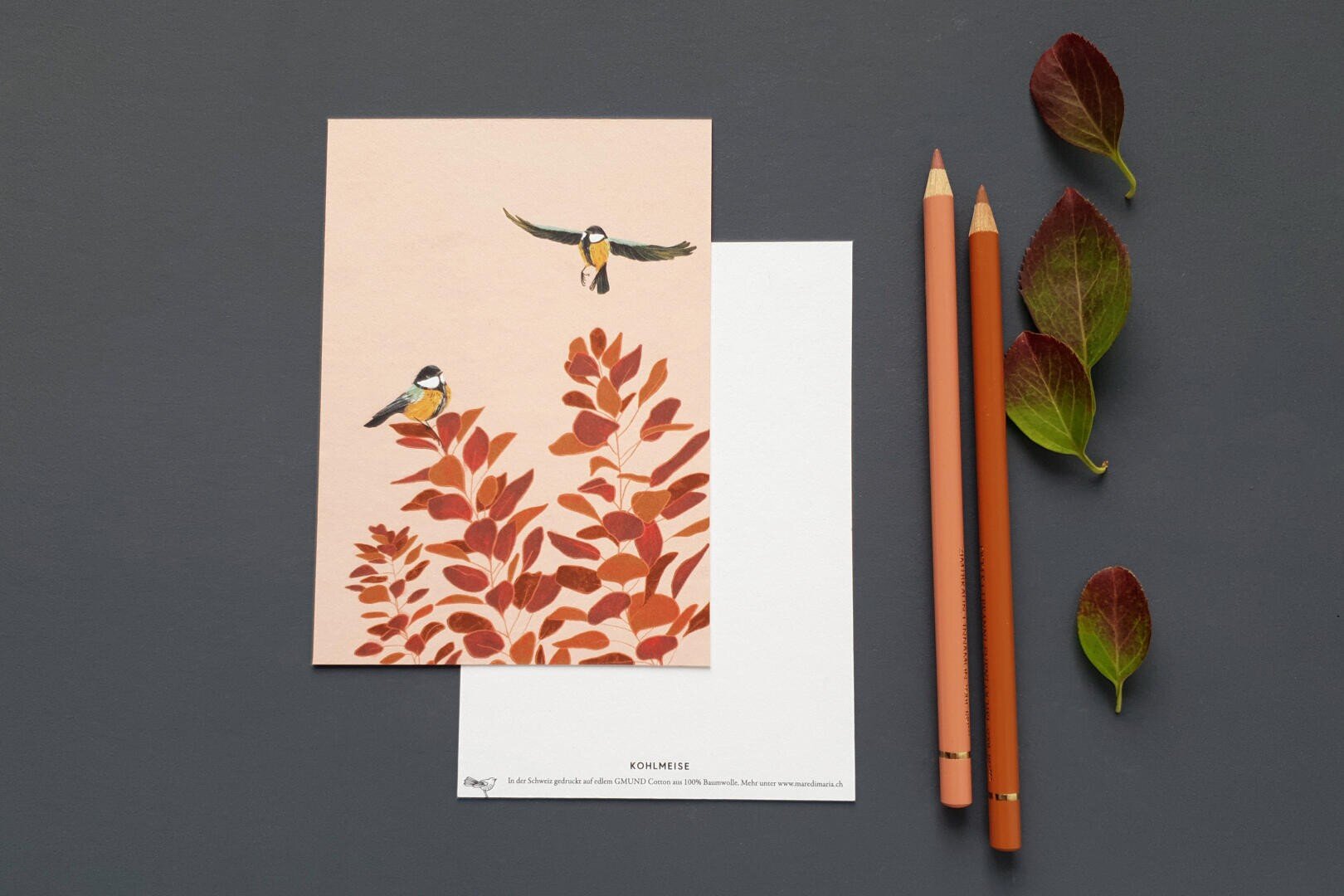 A pink card with two birds and red-orange leaves is placed on a white card. Two orange pencils and three red-green leaves are neatly arranged next to it on a dark gray surface.