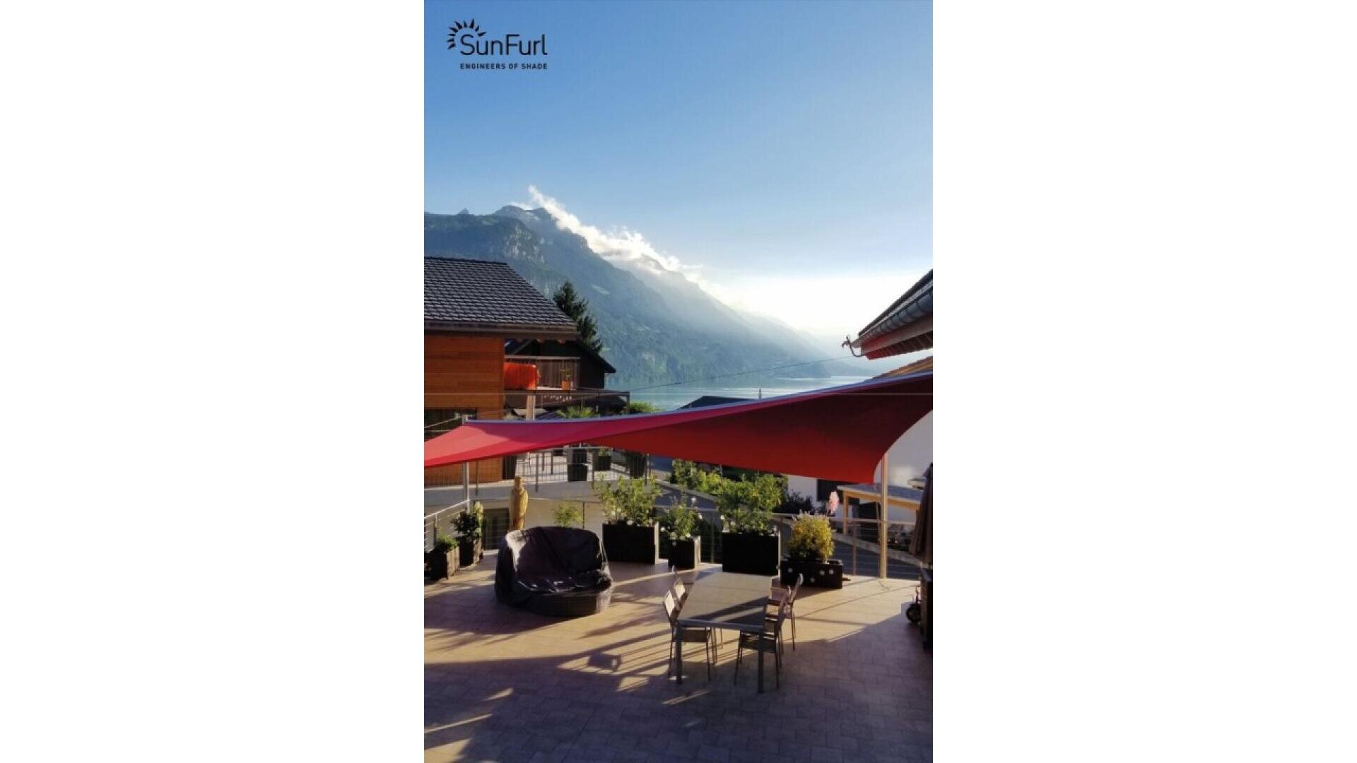 A sunny patio with tables, chairs, and red shade sails overlooks a scenic valley with mountains and a clear blue sky in the background. The SunFurl logo is visible in the top left corner.