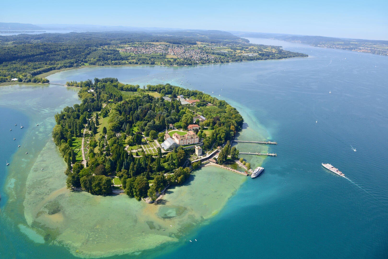 Luftaufnahme einer üppigen, grünen Insel mit Gebäuden und Docks, umgeben von klarem, blauem Wasser. Ein Boot fährt in der Nähe, und das Festland mit Städten und Wäldern ist im Hintergrund unter einem hellen Himmel zu sehen.