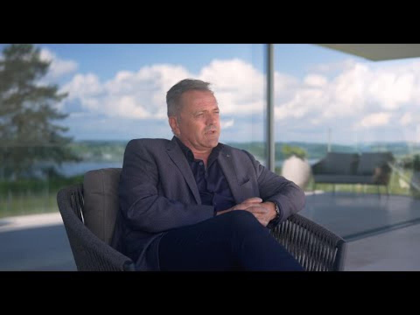 Ein Mann in einem blauen Anzug sitzt auf einem Korbstuhl auf einer Terrasse mit Glasgeländer, mit einer malerischen Aussicht auf Bäume, Wasser und einen bewölkten Himmel im Hintergrund.