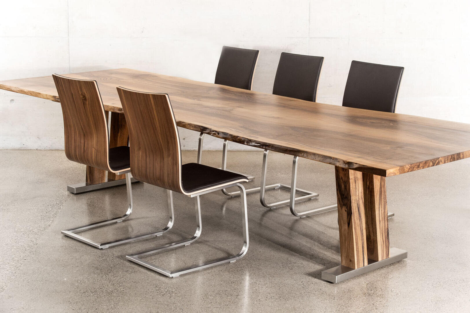 Ein moderner Esstisch aus poliertem Holz mit natürlicher Kante, begleitet von sechs zeitgenössischen Stühlen mit dunkler Sitzfläche und Rückenlehne und verchromten Freischwingergestellen, die auf einem glatten Betonboden stehen.
