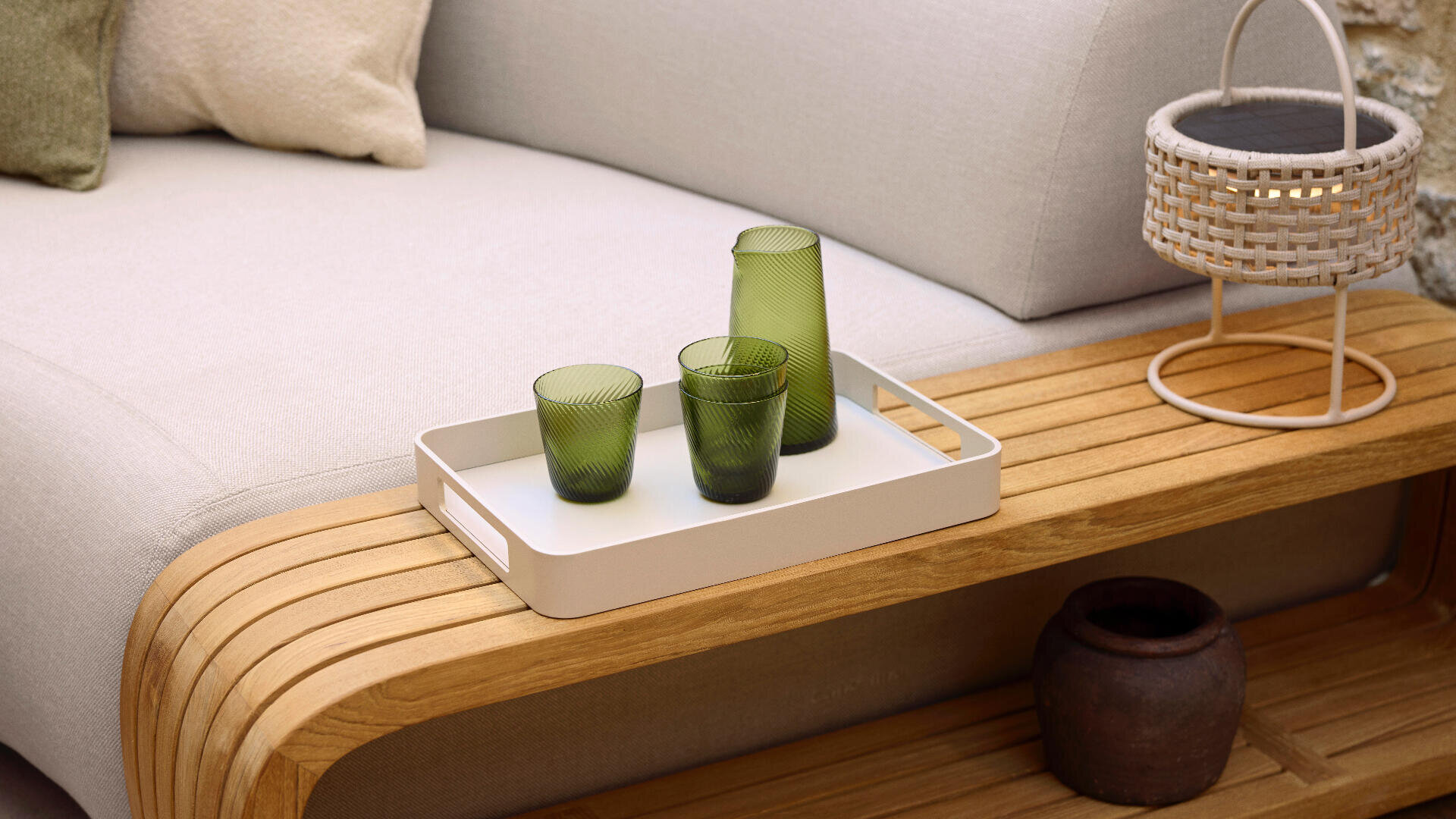 A white tray with two green glasses and a matching green carafe sits on a wooden bench beside a light gray sofa. Nearby are a woven basket and a small dark brown pot.