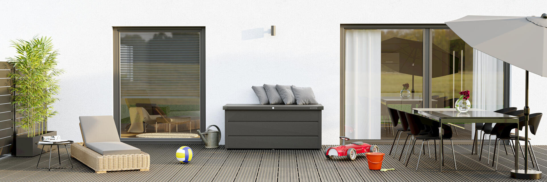Moderne Terrasse mit Korbsessel, Aufbewahrungsbox mit Kissen, Kinderspielzeug, Esstisch mit schwarzen Stühlen, Sonnenschirm, Topfpflanze und großen Glasschiebetüren vor einer weißen Wand.