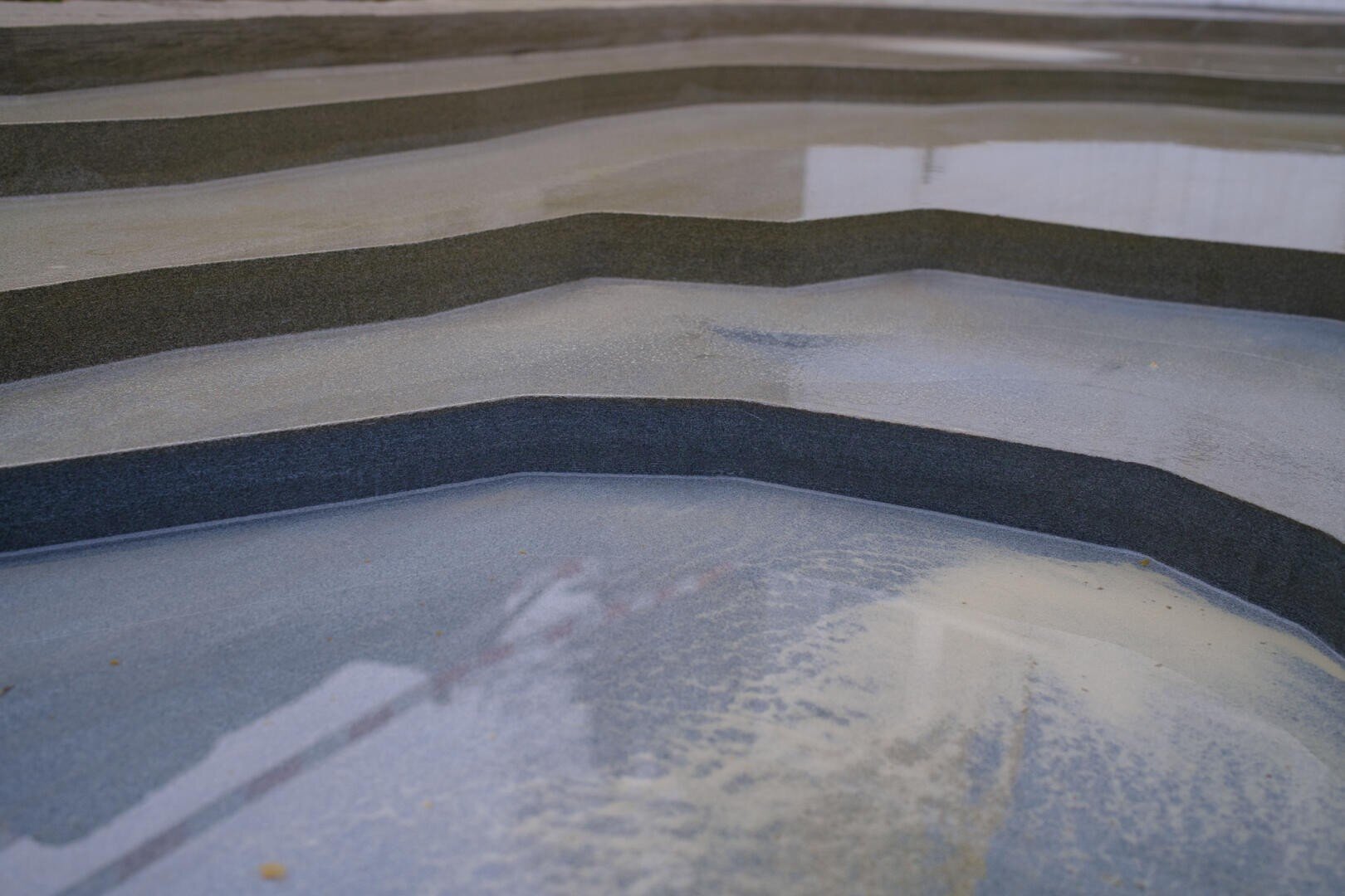 Eine Nahaufnahme von gestuften, gewellten Stein- oder Betonstufen mit flachen Wasserbecken auf jeder Stufe, die einen reflektierenden, geschichteten Effekt erzeugen.