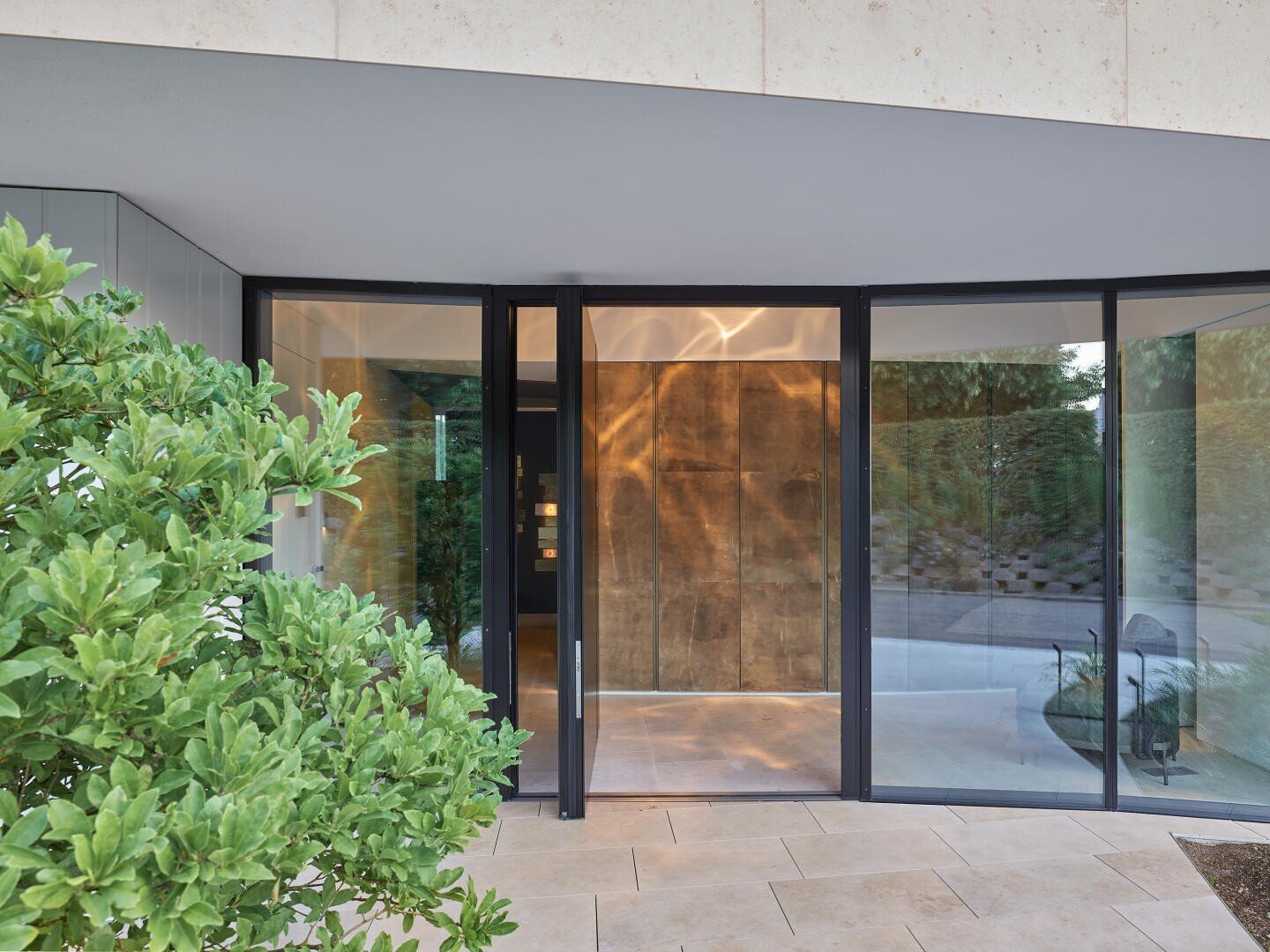 A modern building entrance with large glass doors and windows, light-colored stone flooring and a green leafy plant in the foreground. The interior features warm wood paneling and soft lighting.